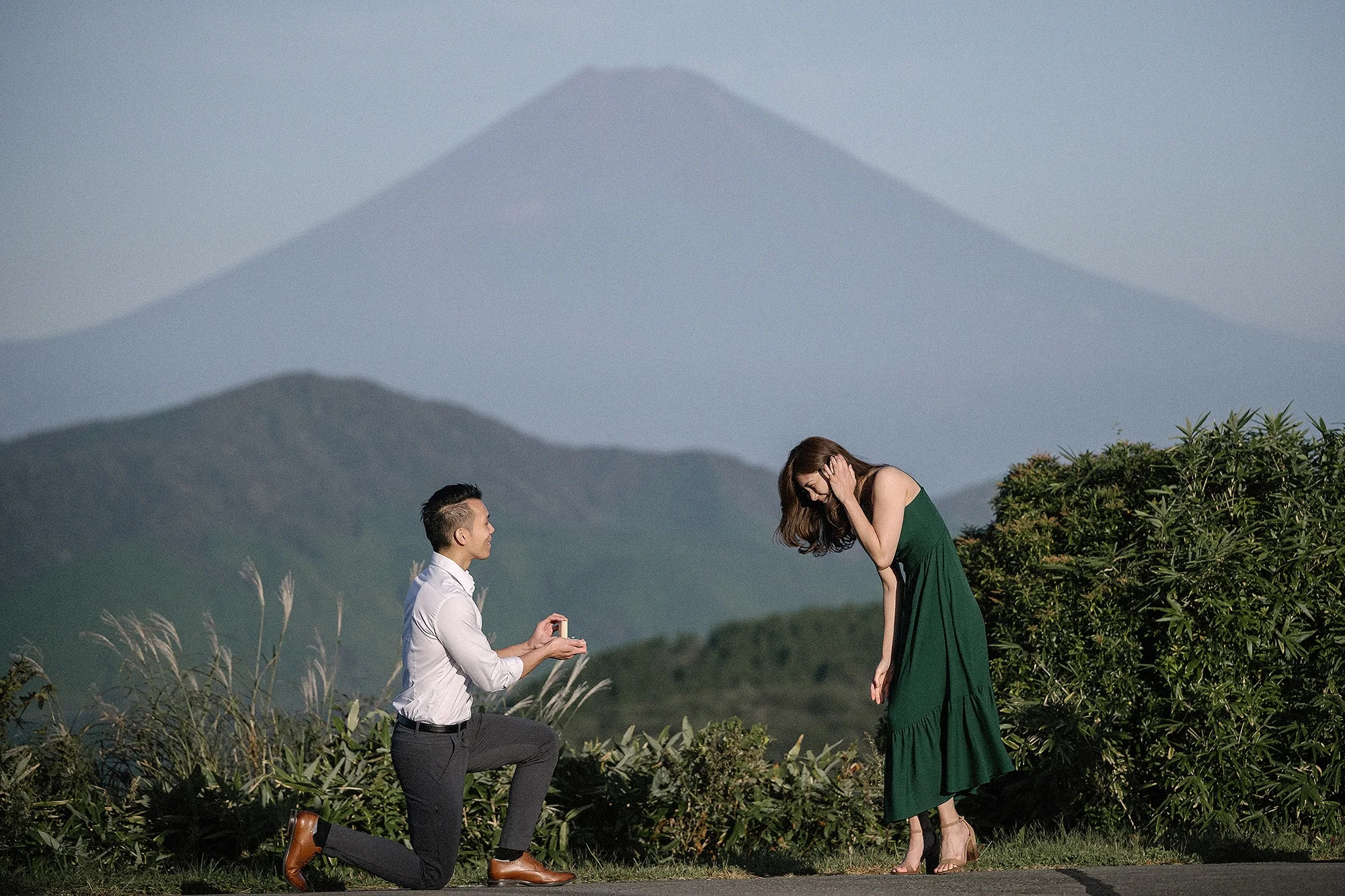 He kneels with a ring on a hilltop above Lake Ashi as she covers her face, Mt Fuji behind them.