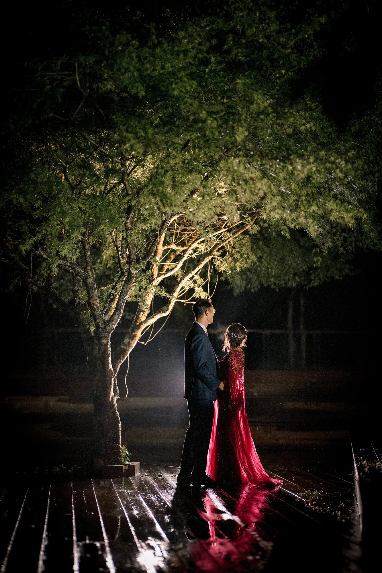 The couple stand together beneath a lit tree at night in Hakone, her red evening gown reflected on the wet deck.