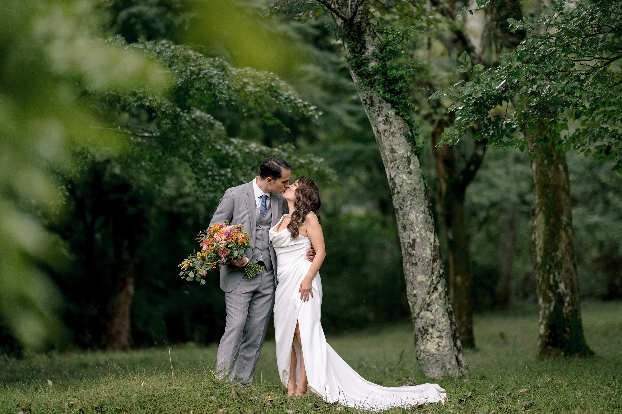 The groom kisses the bride beneath the trees of a Hakone forest, her bouquet of orange and pink flowers in hand.