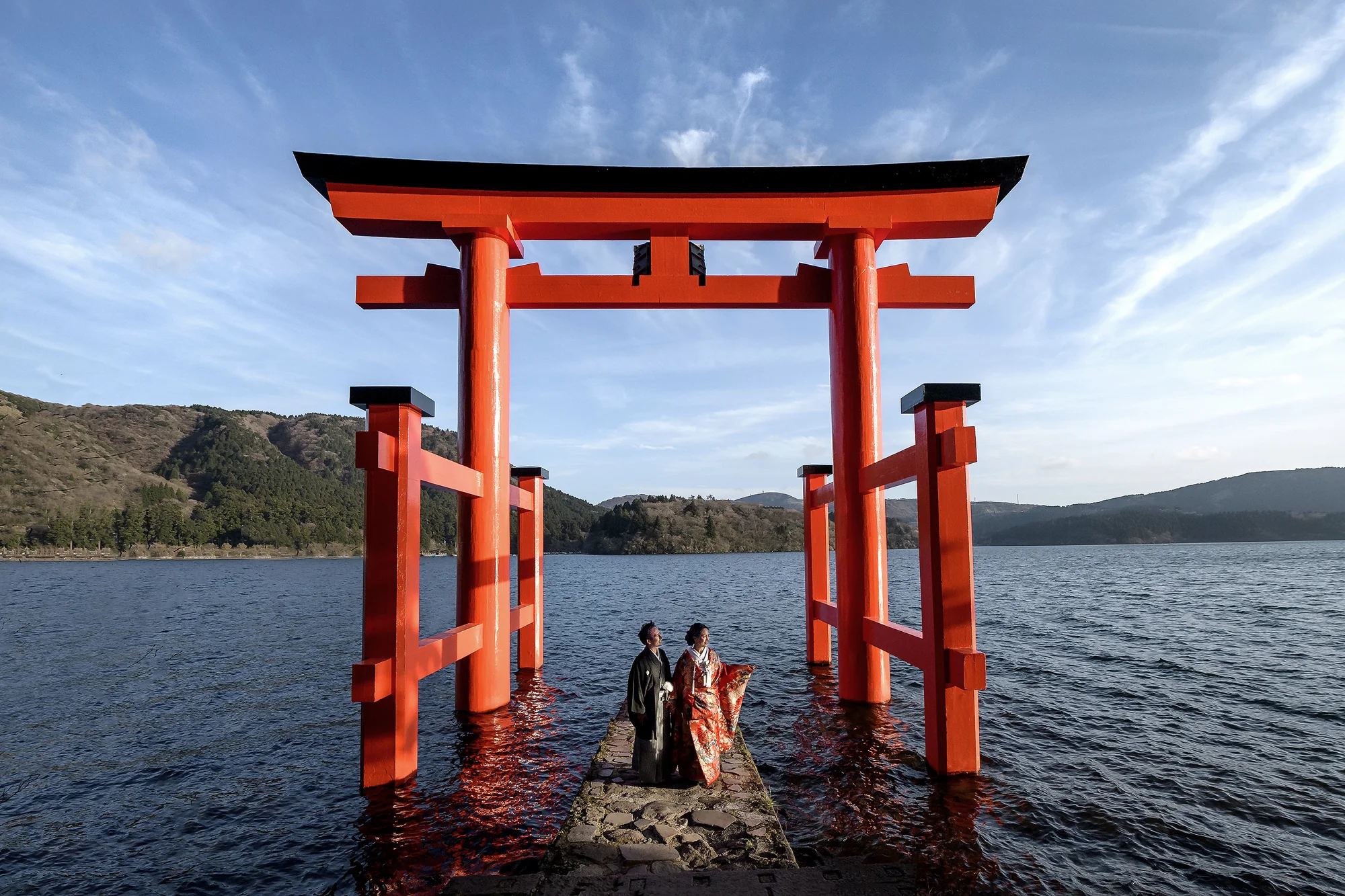 A couple in kimono stand on the stone steps of a vermilion torii rising from Lake Ashi in Hakone.