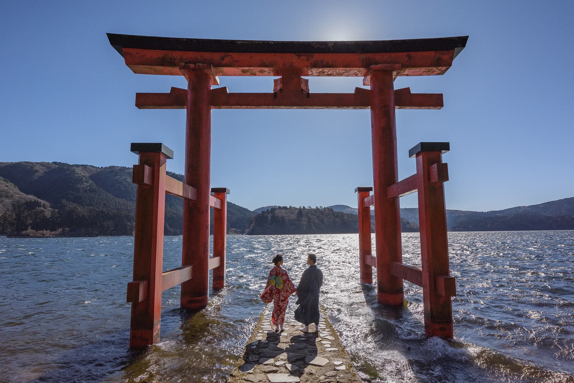 A couple in kimono stand on the stone steps of a vermilion torii in Lake Ashi under a clear blue sky in Hakone.