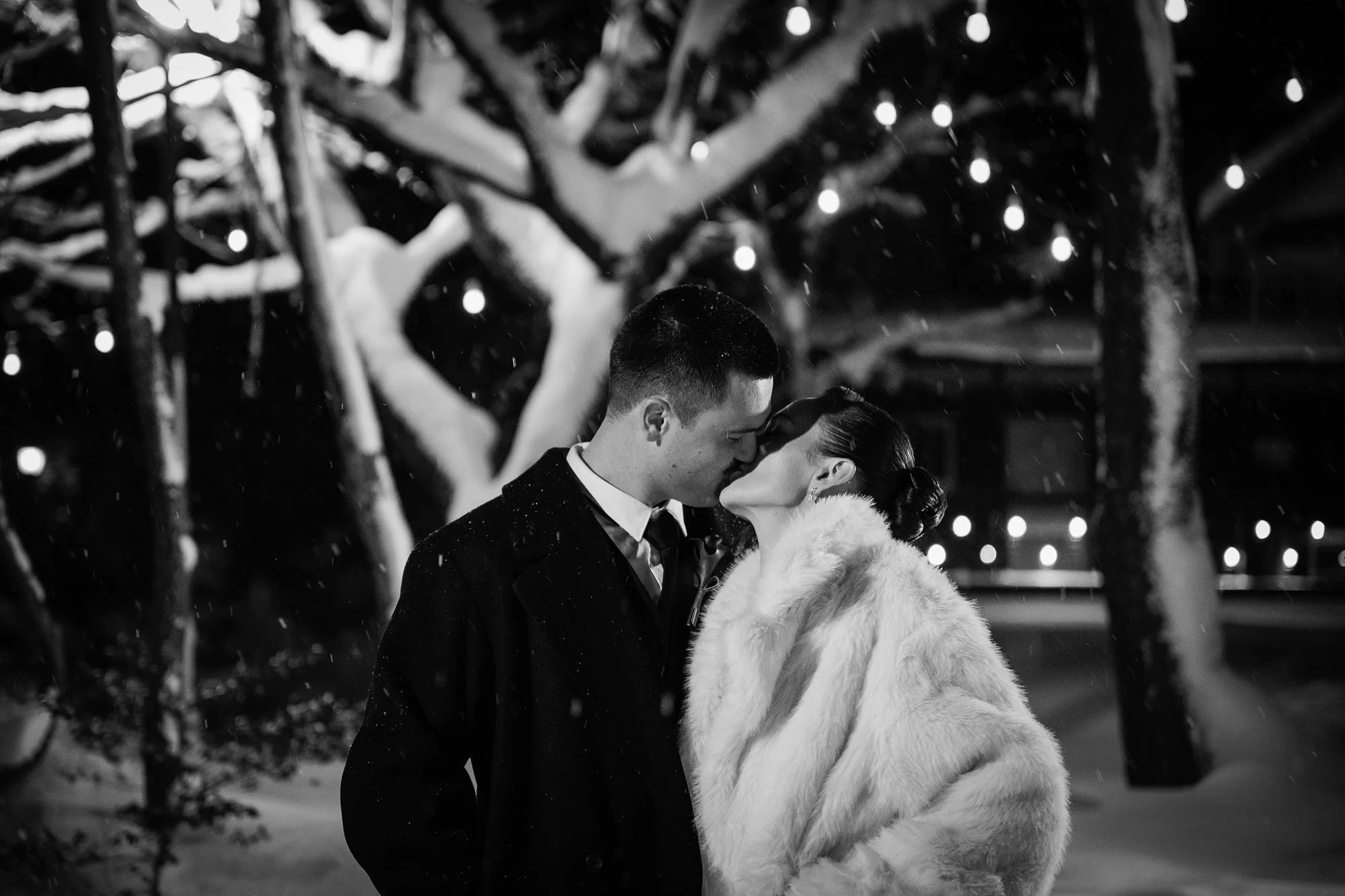 In black and white, the couple kiss at night as snow falls among lit trees.