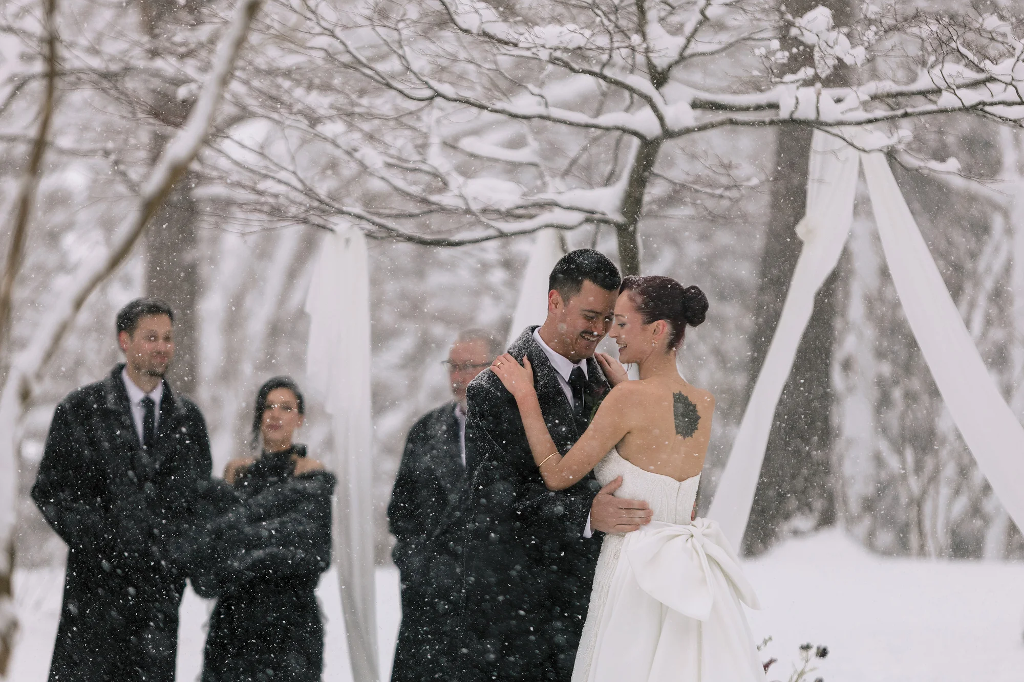 The bride turns, laughing, as the groom smiles at her during their ceremony in falling snow, guests in dark coats around them.
