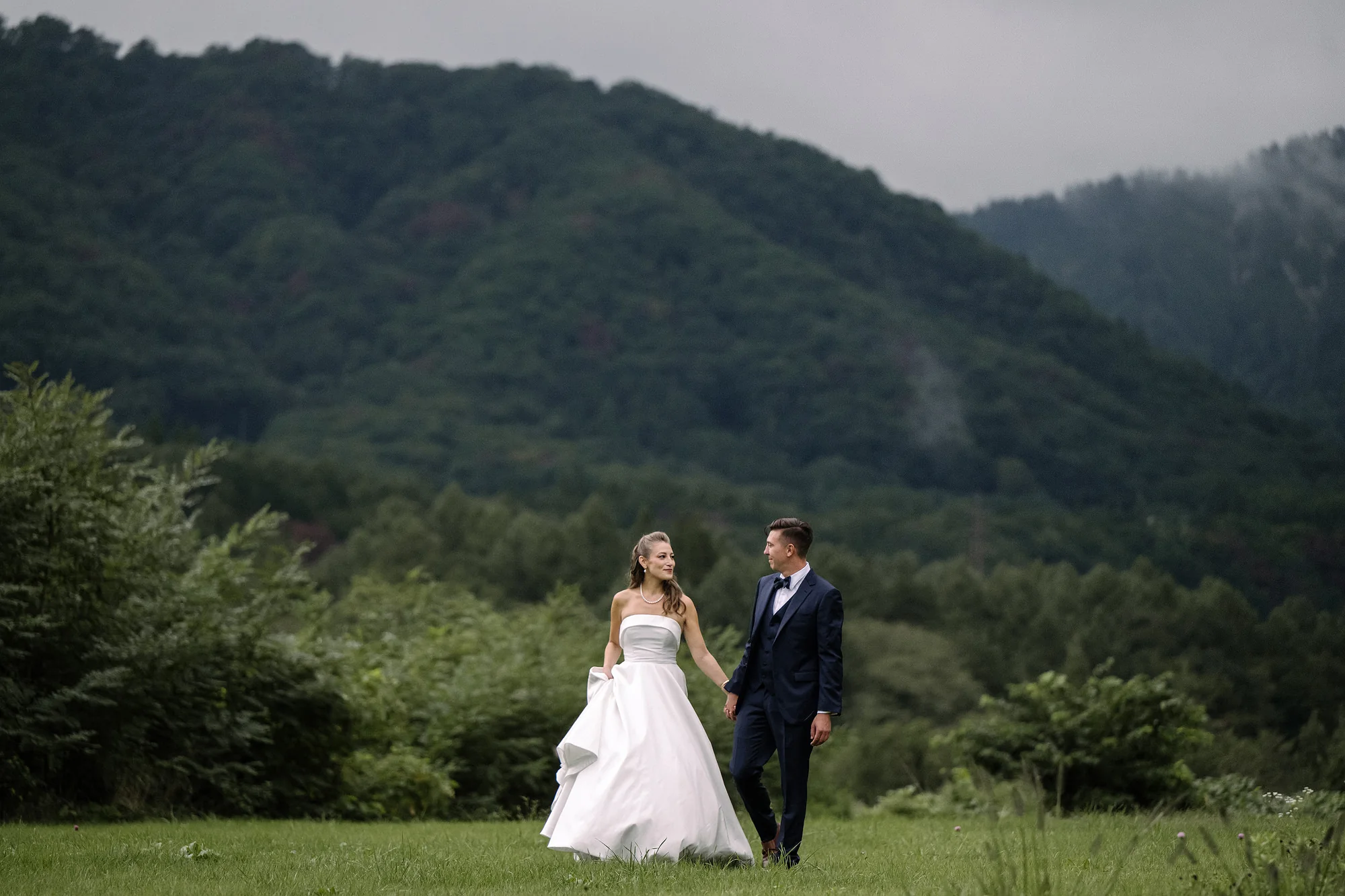 A couple walk across a green meadow in a Hakuba valley in summer, mist on the slopes behind.