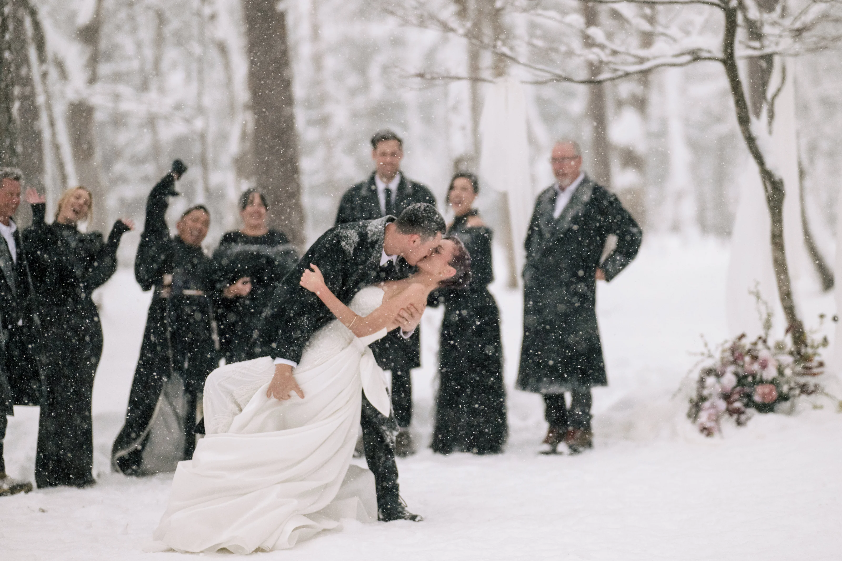 The bride and groom kiss in the falling snow as their wedding guests celebrate behind them in a Hakuba forest.