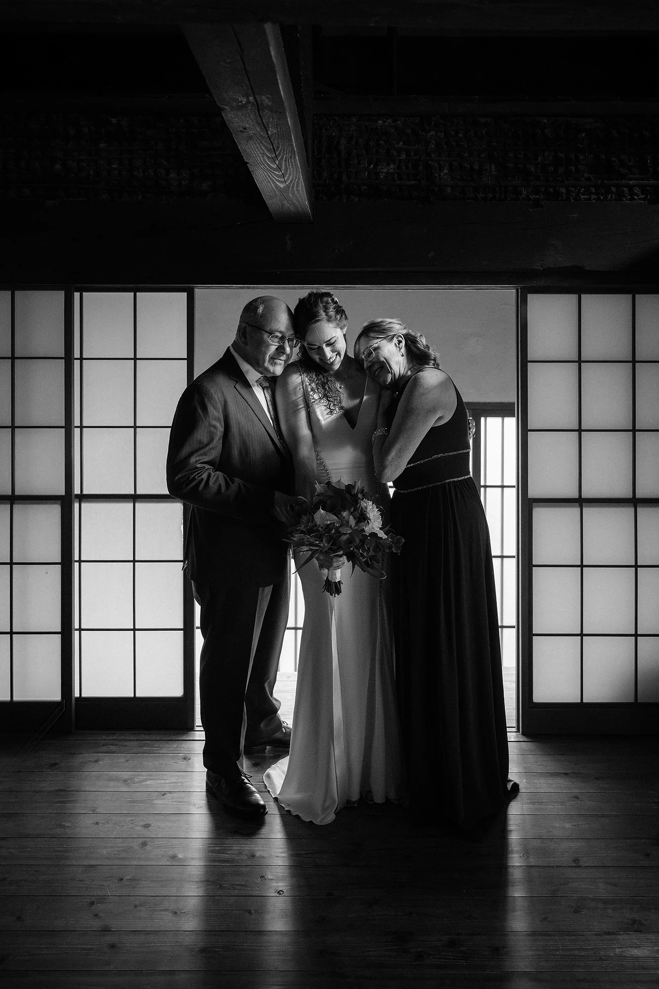 In black and white, the bride stands between her parents, heads close together, in a timber room lit by tall shoji windows at NIPPONIA Kosuge.