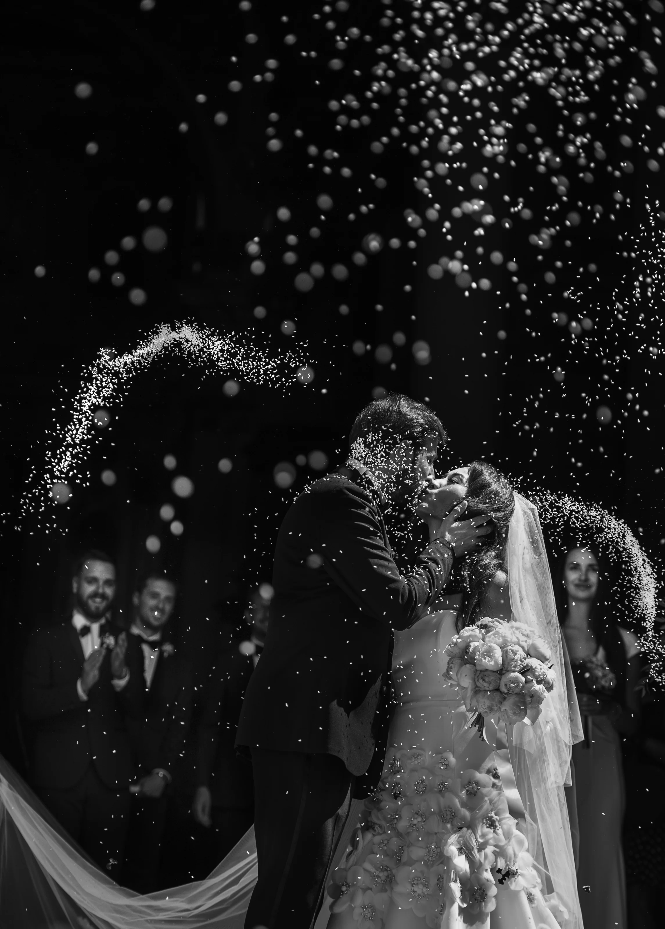 In black and white, the couple kiss as guests throw confetti that glitters in the light, in Italy.