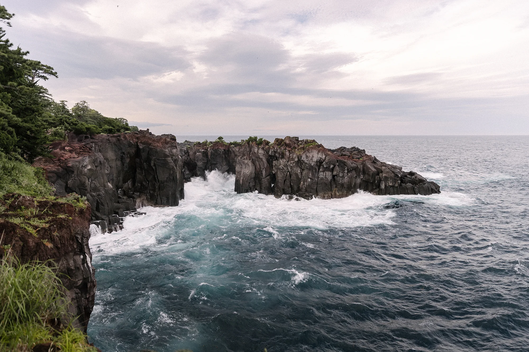 Waves break against the dark volcanic cliffs of the Izu coast under a pale sky.