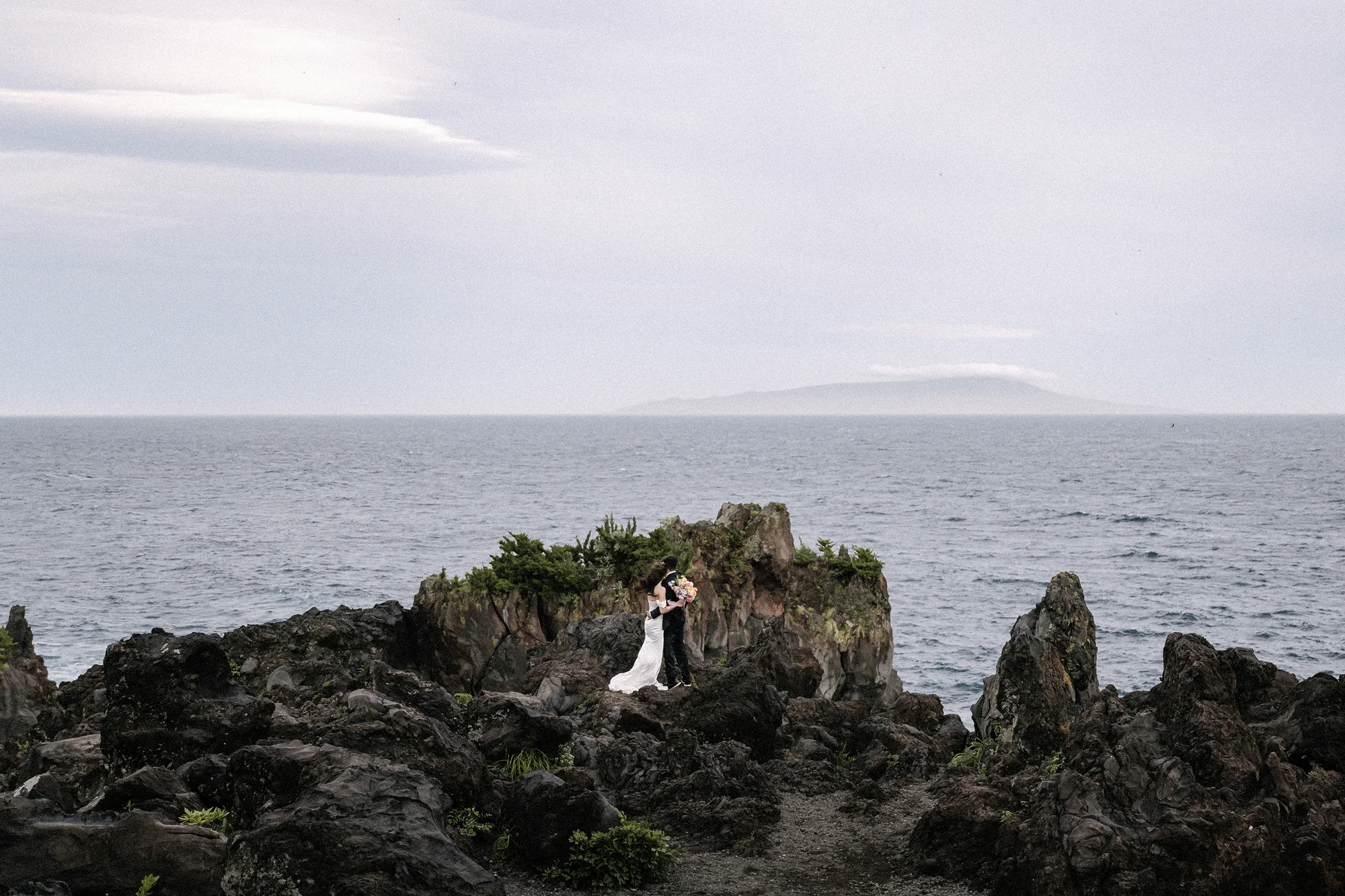 A couple stand small on black volcanic rocks above the sea on the Izu coast, an island faint on the horizon.