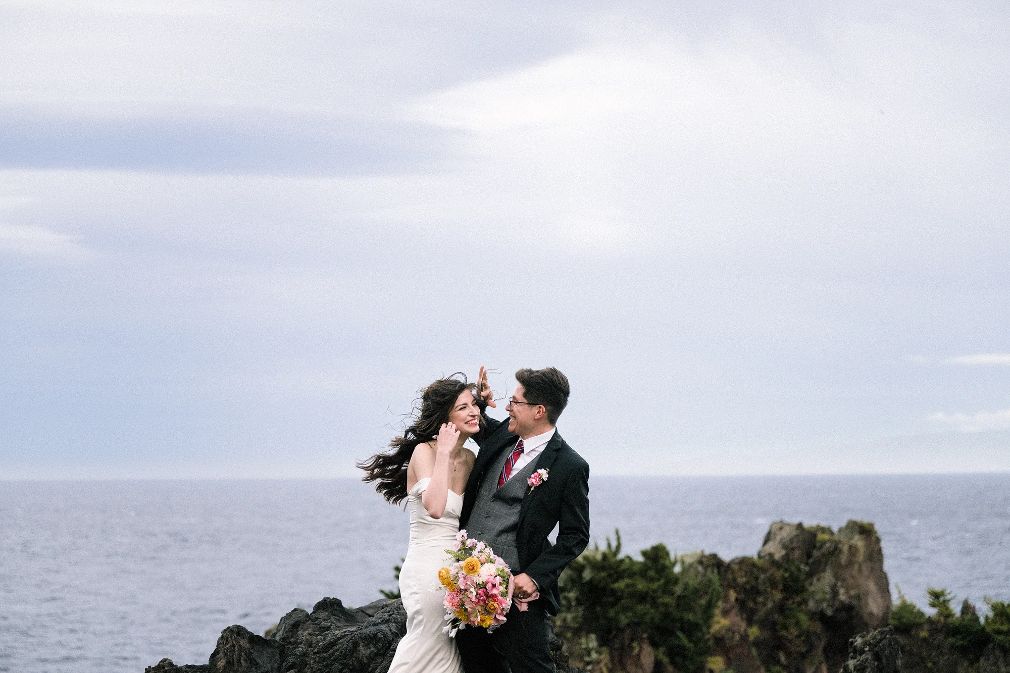 The bride laughs as the wind catches her veil, the groom beside her on a cliff above the sea.