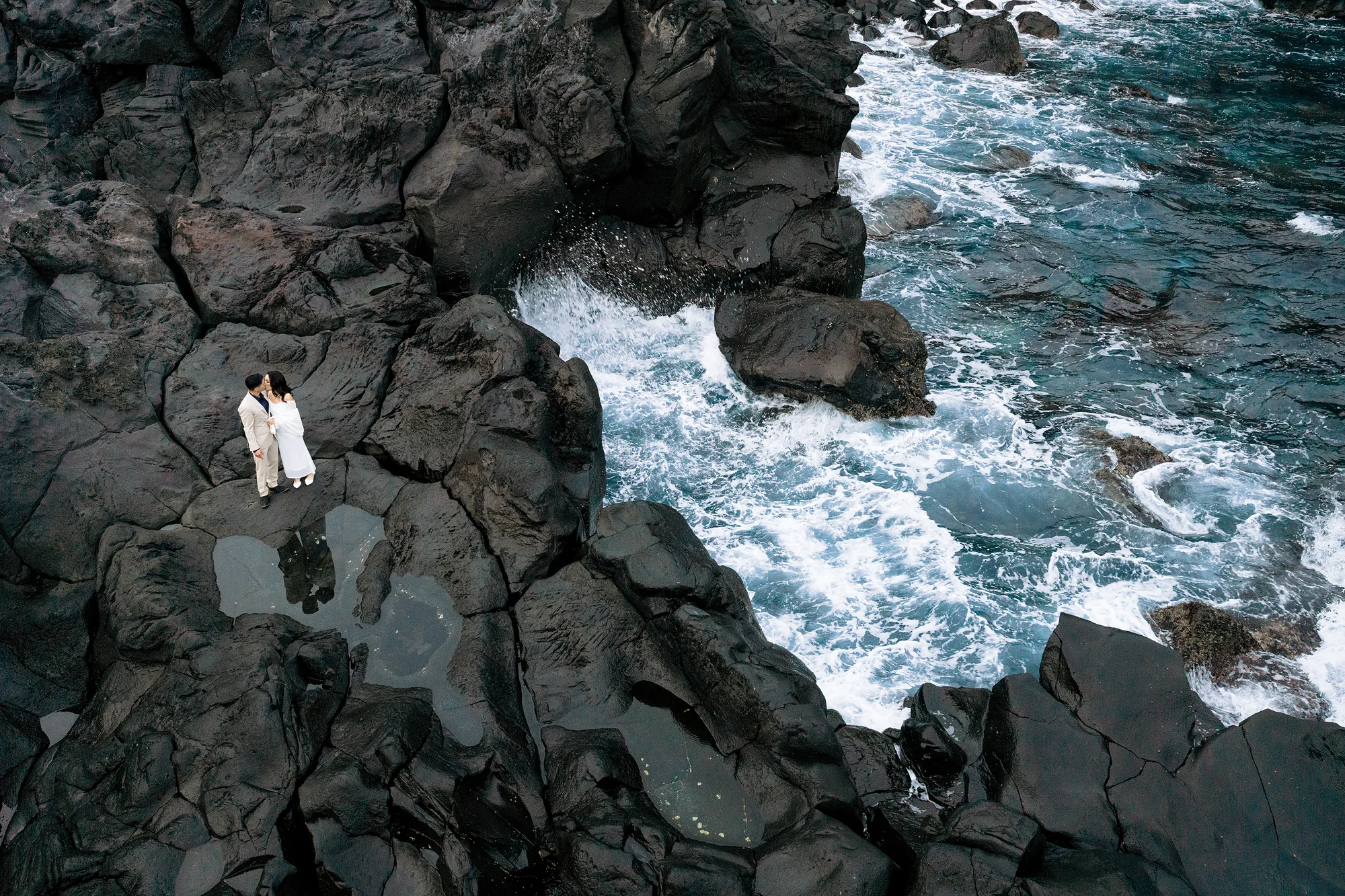 From above, a couple stand on black volcanic rocks as the surf breaks below them on the Izu coast.