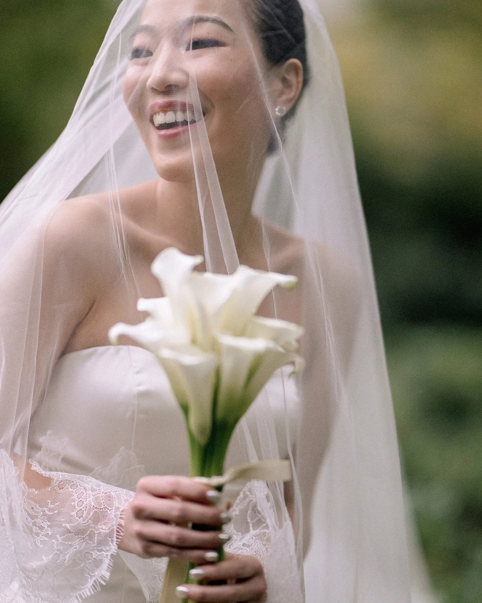 The bride laughs under her veil, holding white calla lilies in the garden at Jinya Ryokan on the day of a Greek and Japanese wedding.