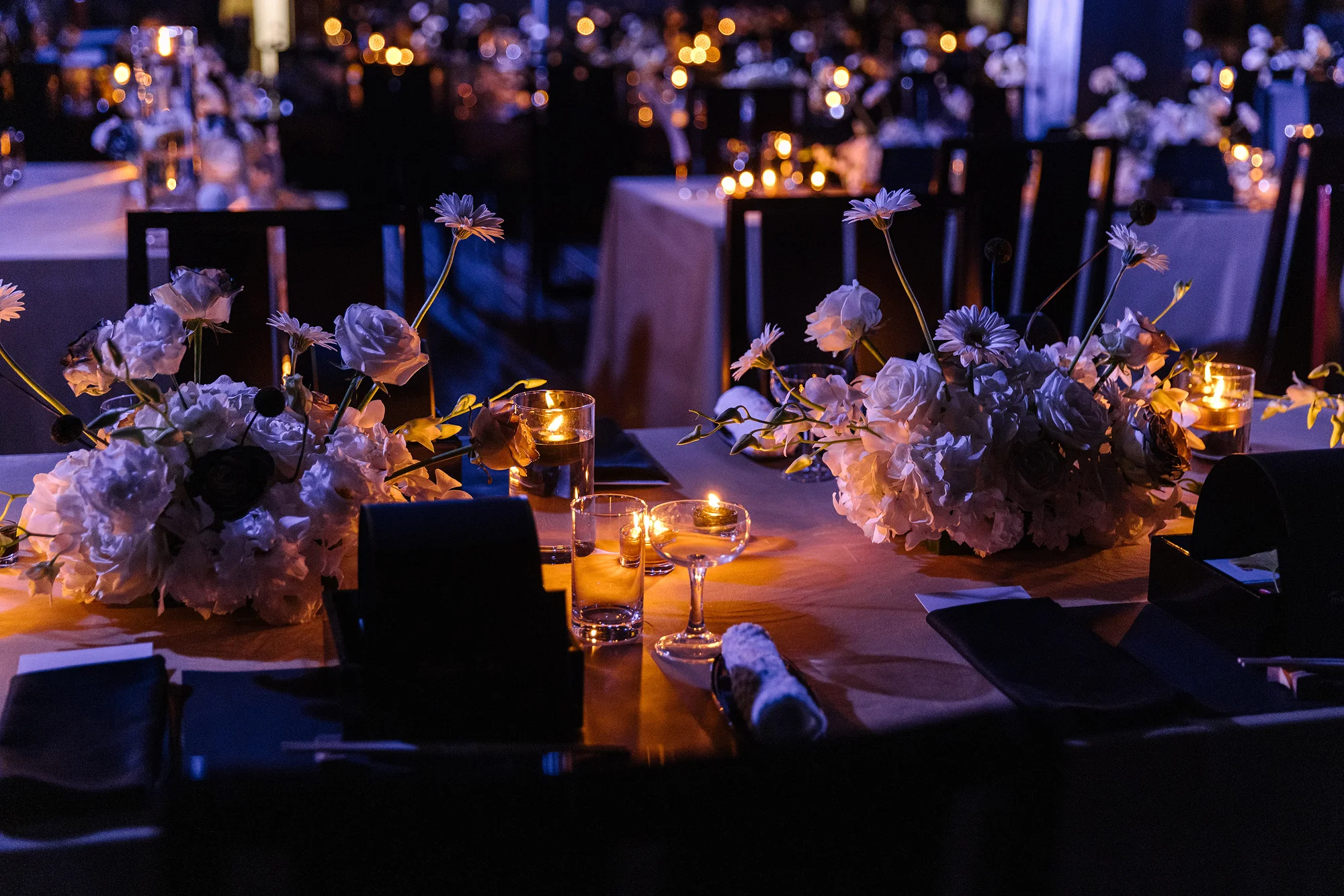 Tealights and floating candles glow among low arrangements of white roses and gerberas on a reception table at night, the room beyond in soft focus.