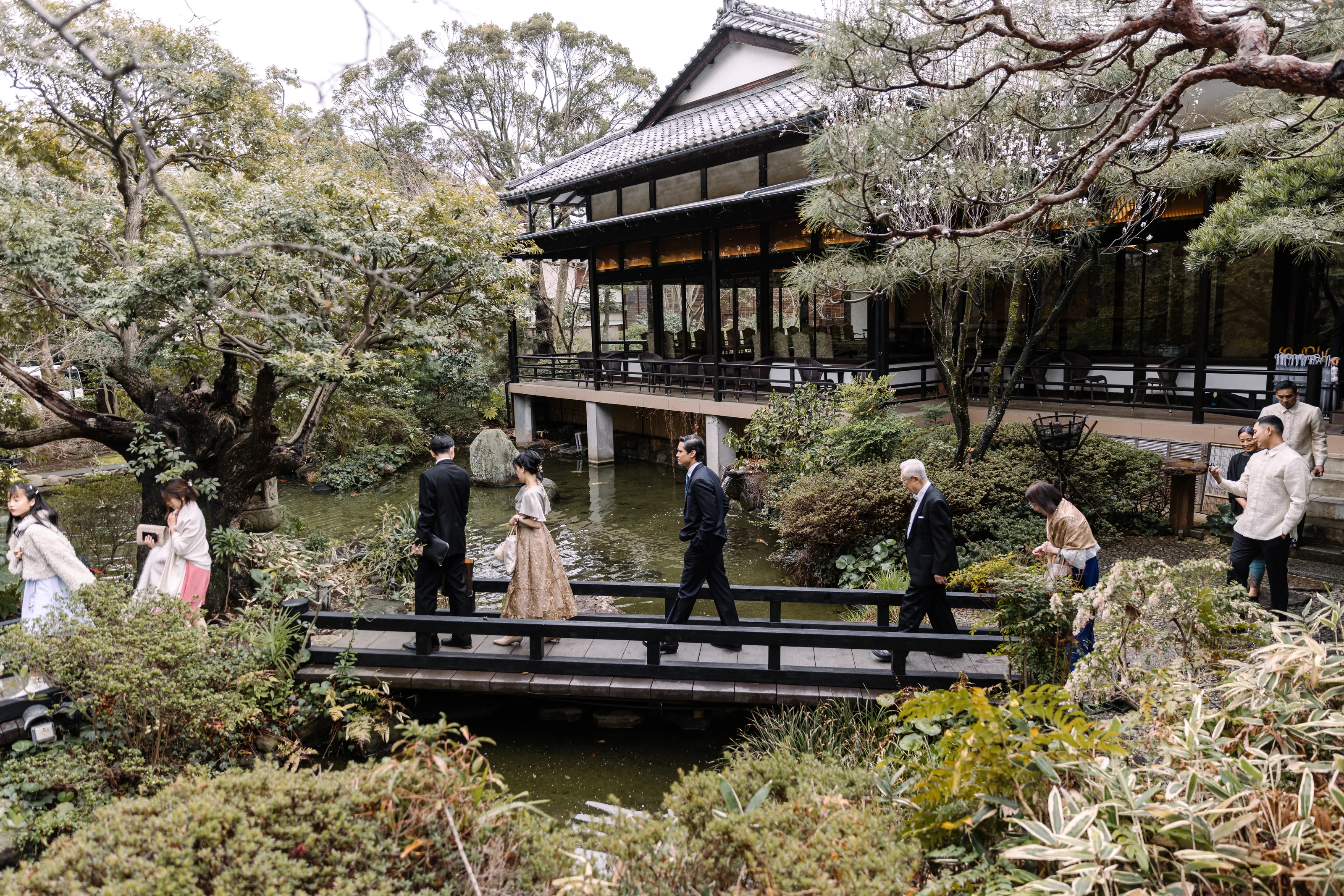 Wedding guests cross a wooden bridge over the pond at Motoyu Jinya, with the old wooden hall and pines of the garden behind.