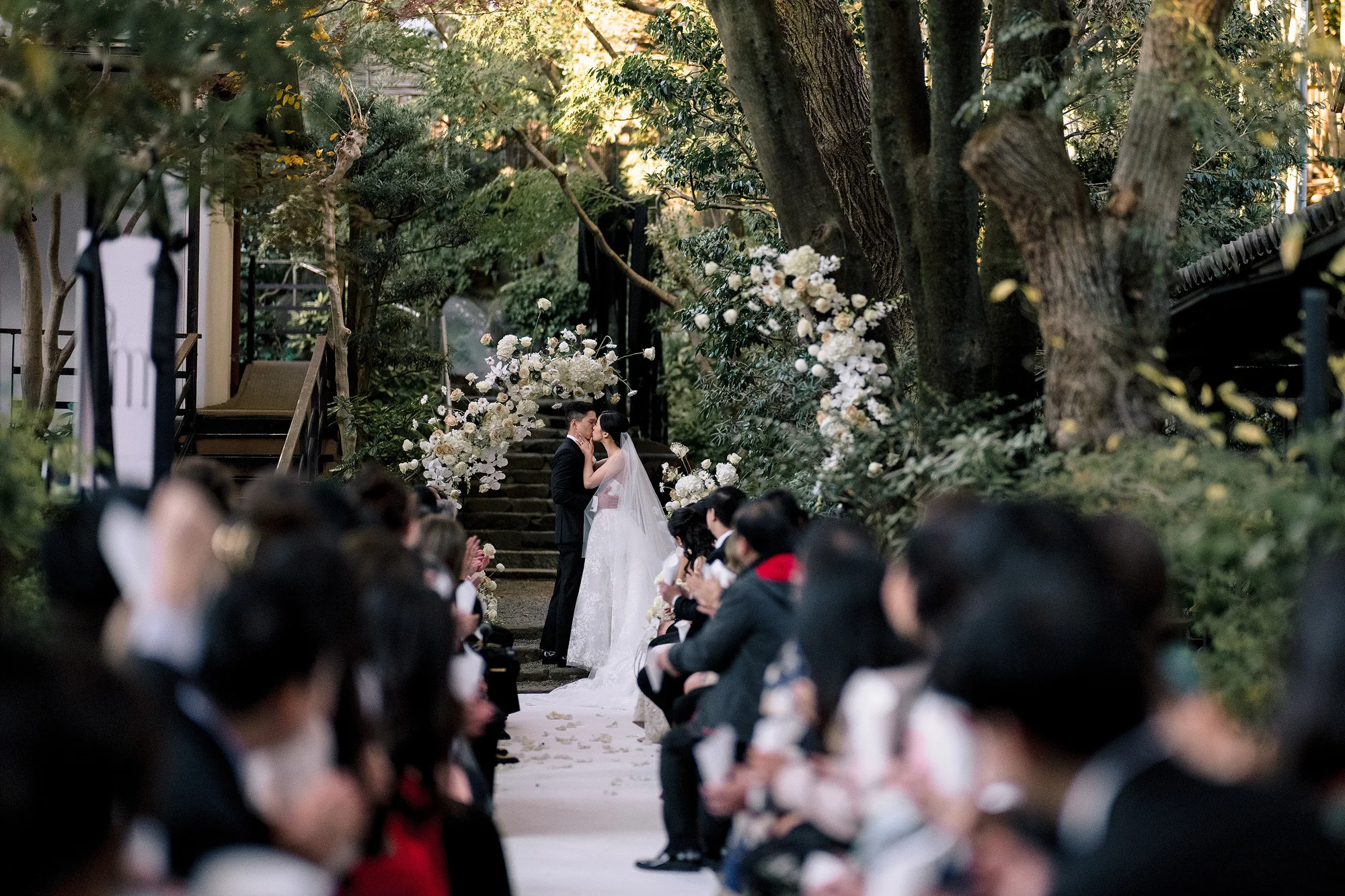 Guests watch from both sides of the aisle as the couple kiss beneath a white floral arch in the garden at Jinya.