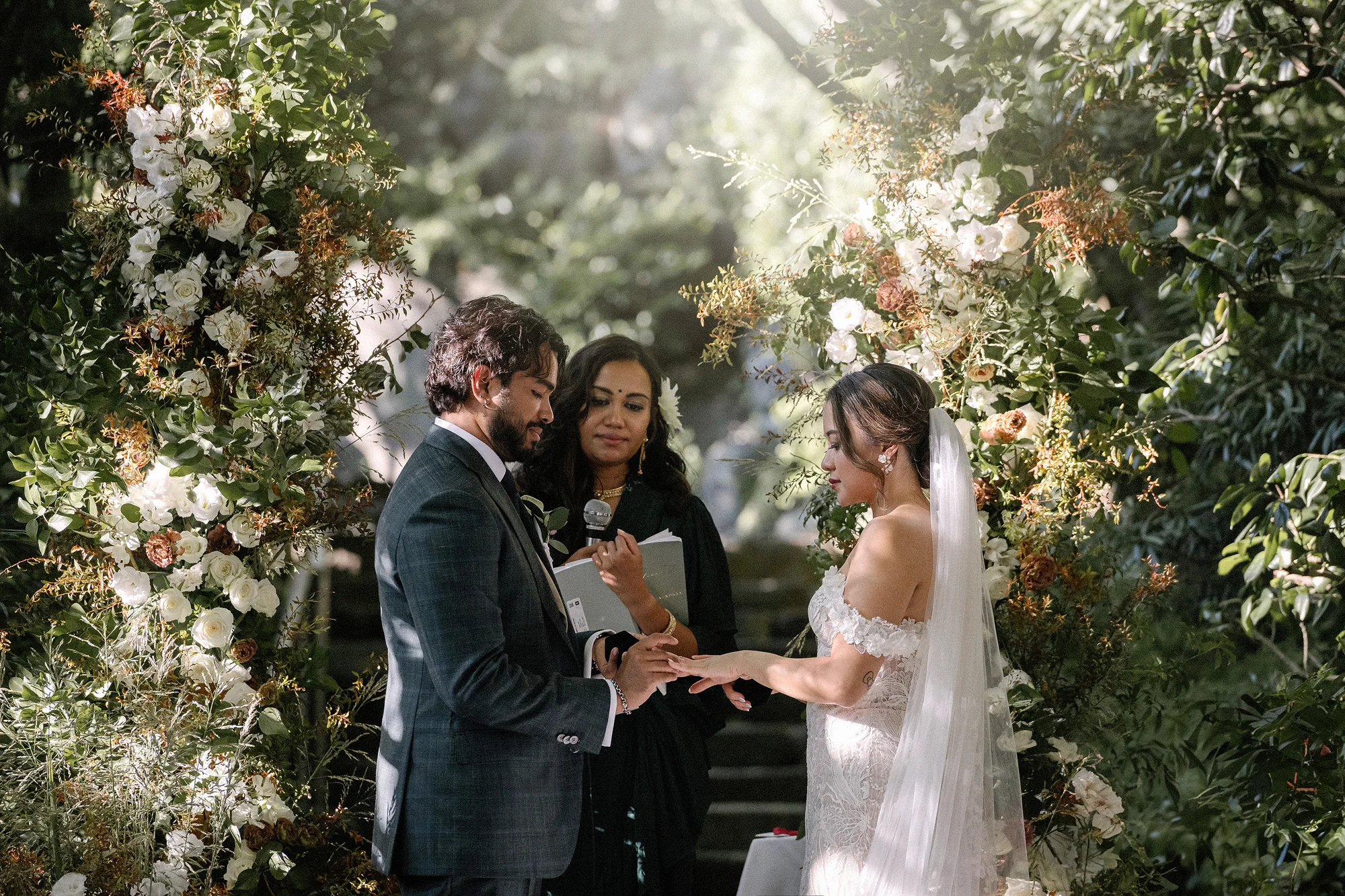 The groom places the ring on the bride's finger beneath a ceremony arch of white roses and autumn leaves among the trees at Jinya.