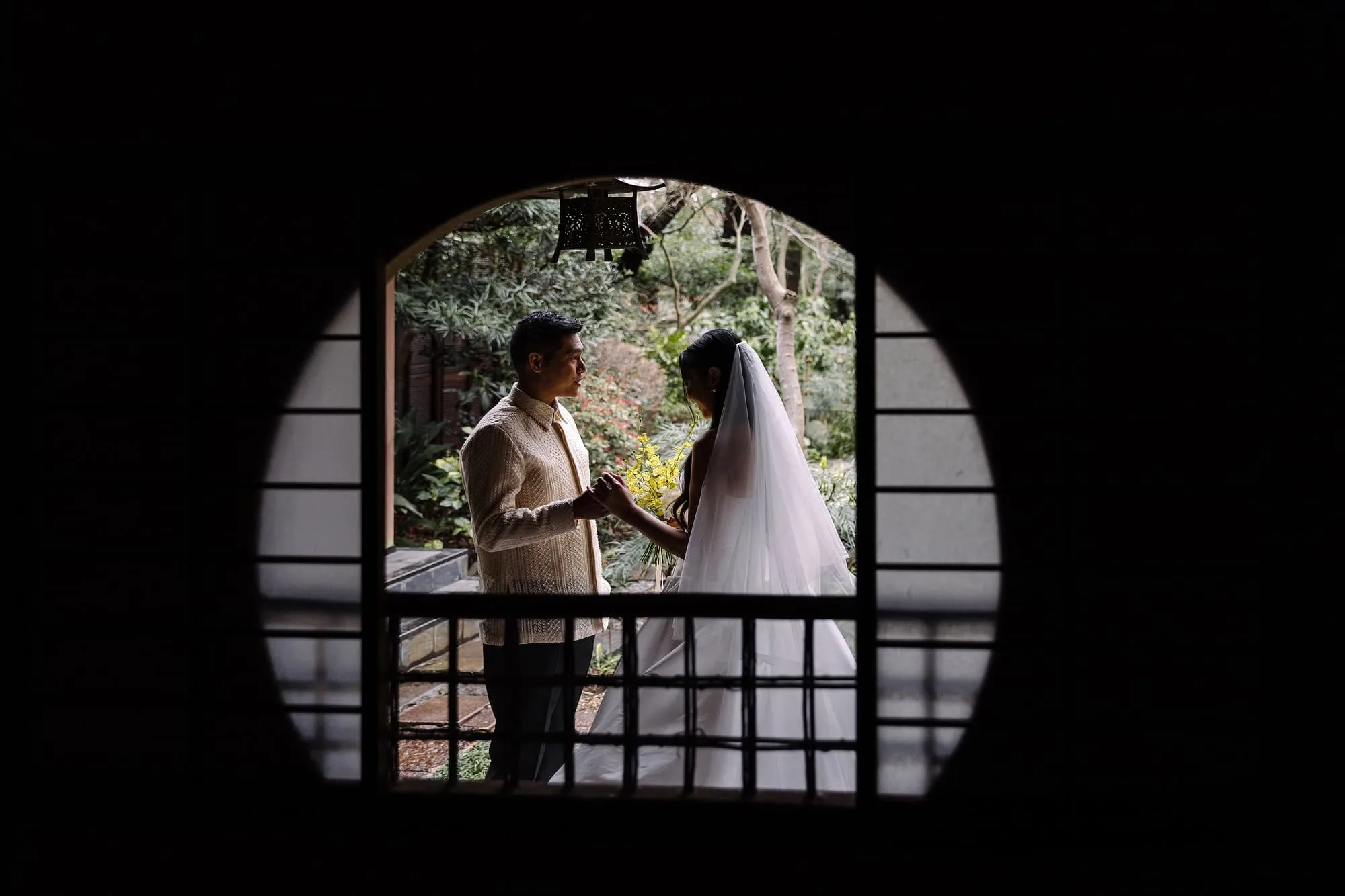 The bride and groom hold hands framed by a round window at their onsen wedding in Japan, a dark wooden room around them.