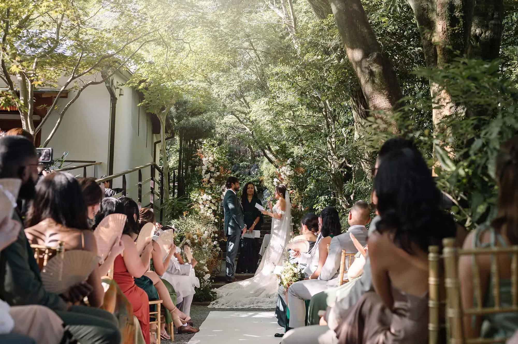 Looking down the walkway aisle at Jinya from the floral arch, guests seated on gold chairs under the trees at an outdoor wedding in Japan.