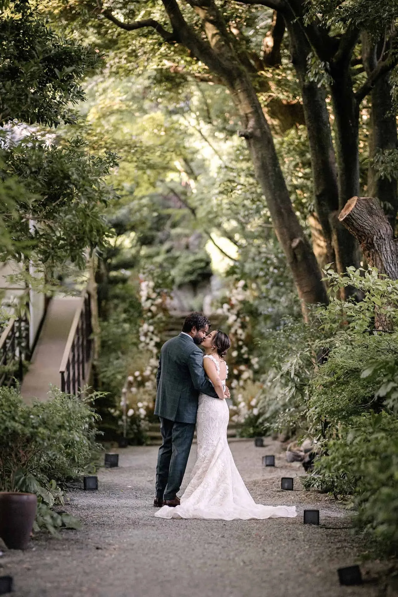 The bride and groom kiss far down a gravel garden path under tall trees at Jinya in Tsurumaki Onsen, Kanagawa.