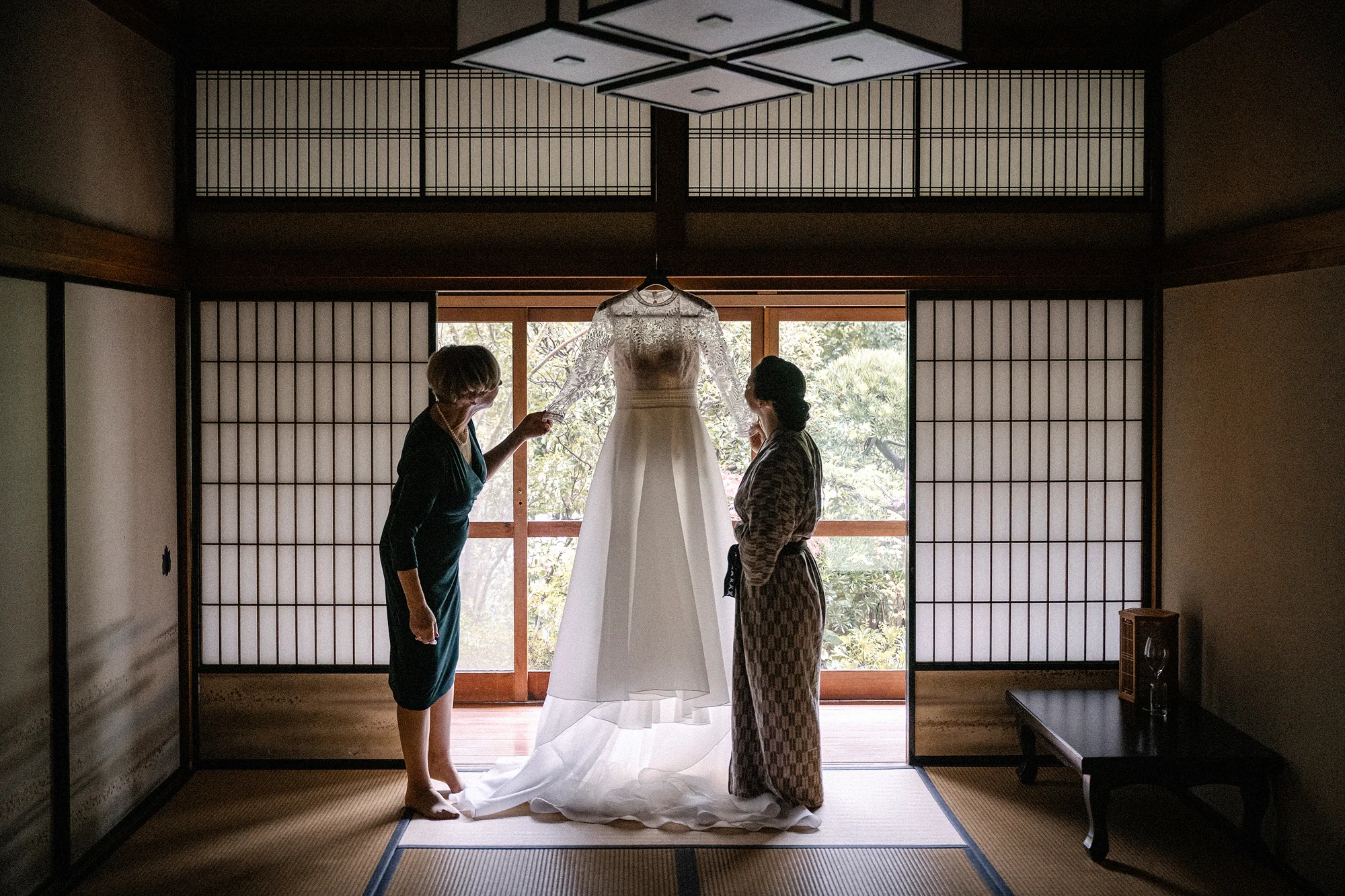 The bride's mother and the bride, in a patterned robe, admire the wedding dress hanging in the doorway of a tatami room, shoji screens on either side.