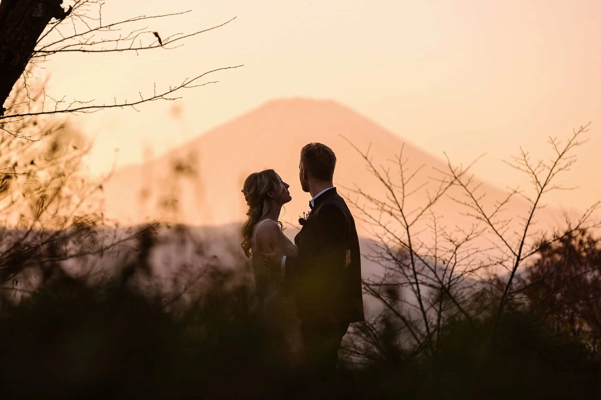 The bride looks up at the groom as he holds her, with Mt Fuji behind them in a pink sunset haze, at the end of an autumn elopement in Japan.