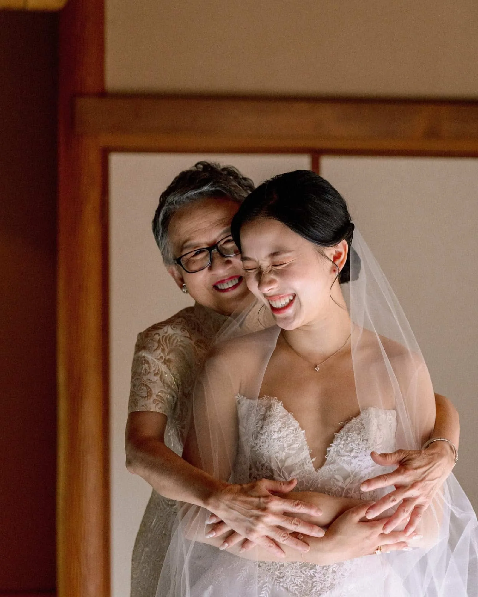 The bride's mother hugs her from behind as they both laugh, veil on, while the bride gets ready in a wood-framed ryokan room at Jinya.