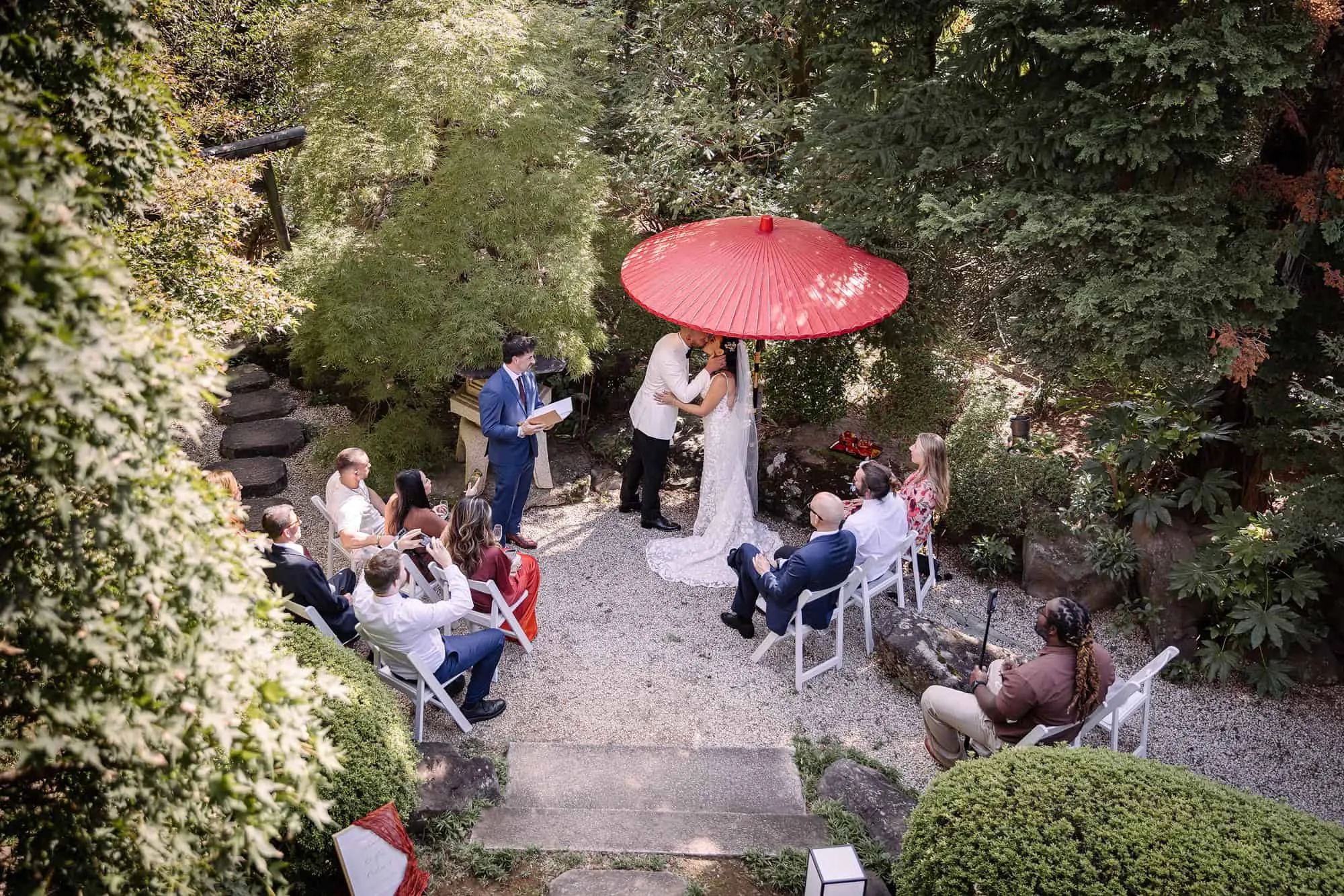 Friends watch from white chairs as the couple kiss beneath a red wagasa in the garden at Tantoku, a Japanese garden wedding in Kawagoe.