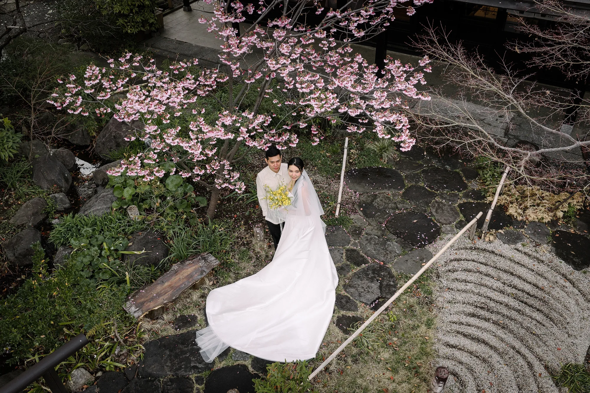 The bride and groom stand beneath pink cherry blossom beside raked gravel in the garden at Motoyu Jinya, her train sweeping the stones.