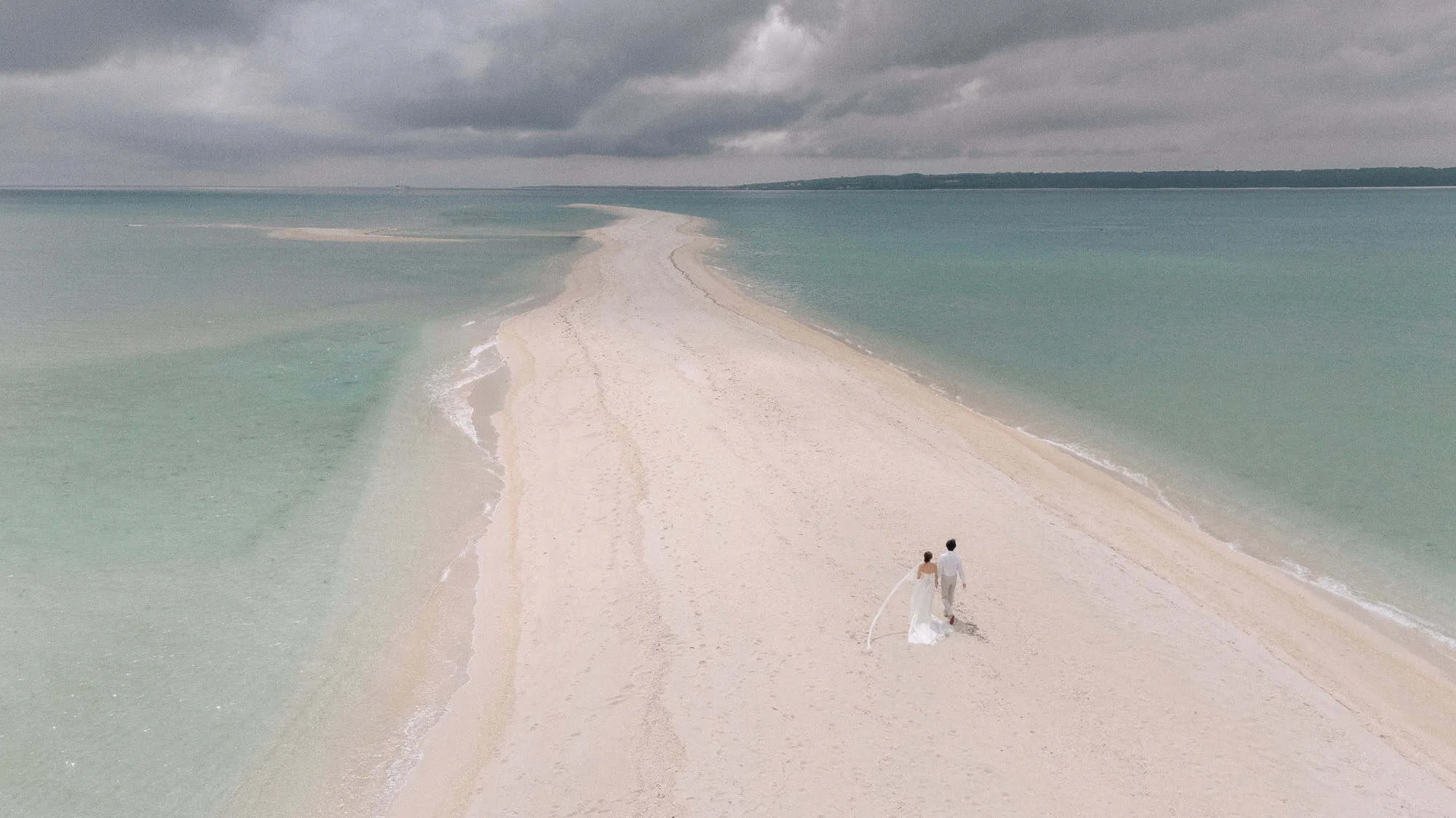 The couple walk a long sandbar through turquoise sea under storm cloud, her veil trailing, the day before their Rosewood Miyakojima wedding.