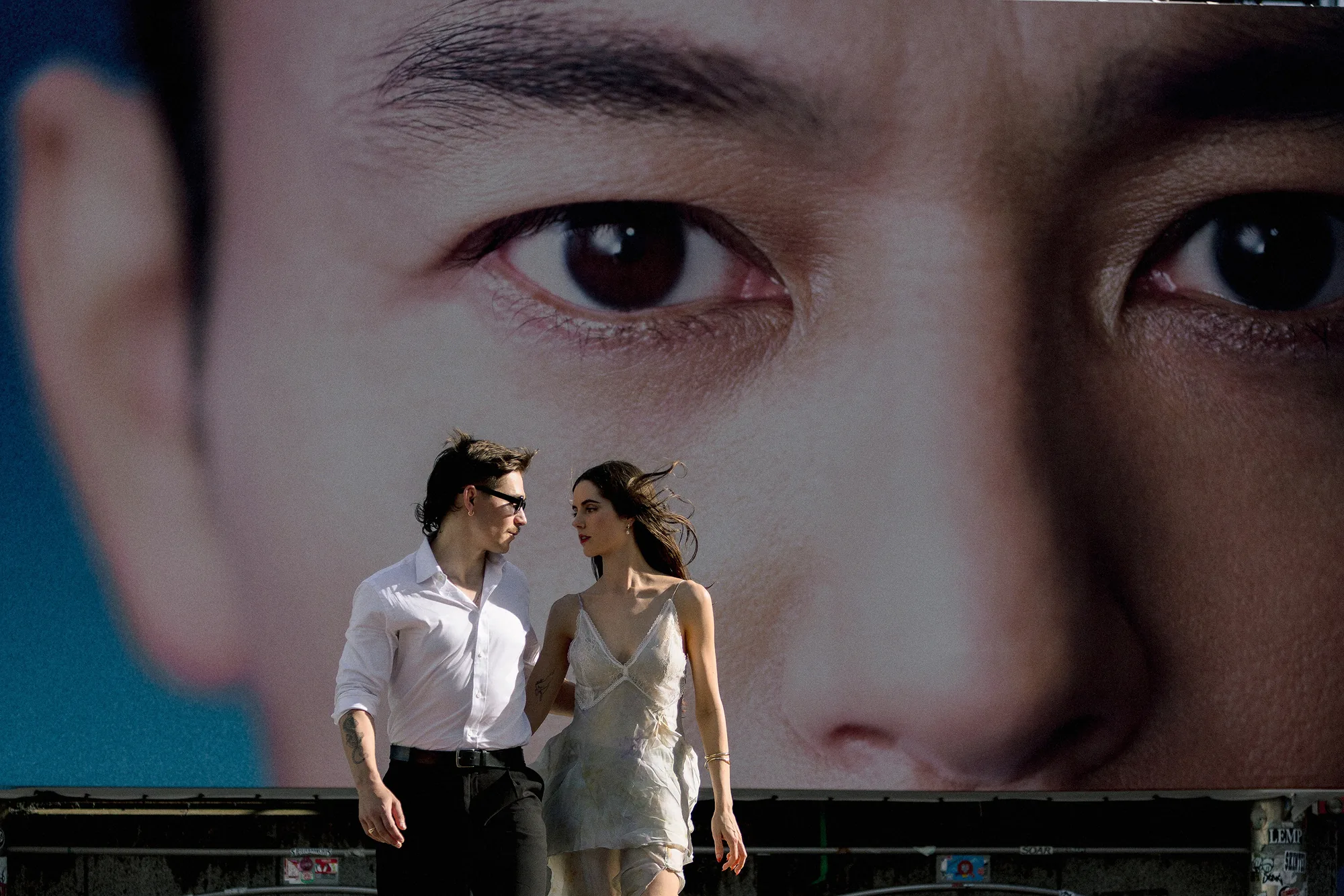 The couple walk past a giant billboard face in Shibuya, its eyes wider than they are tall, in Shibuya pre-wedding photos taken two nights before their wedding.