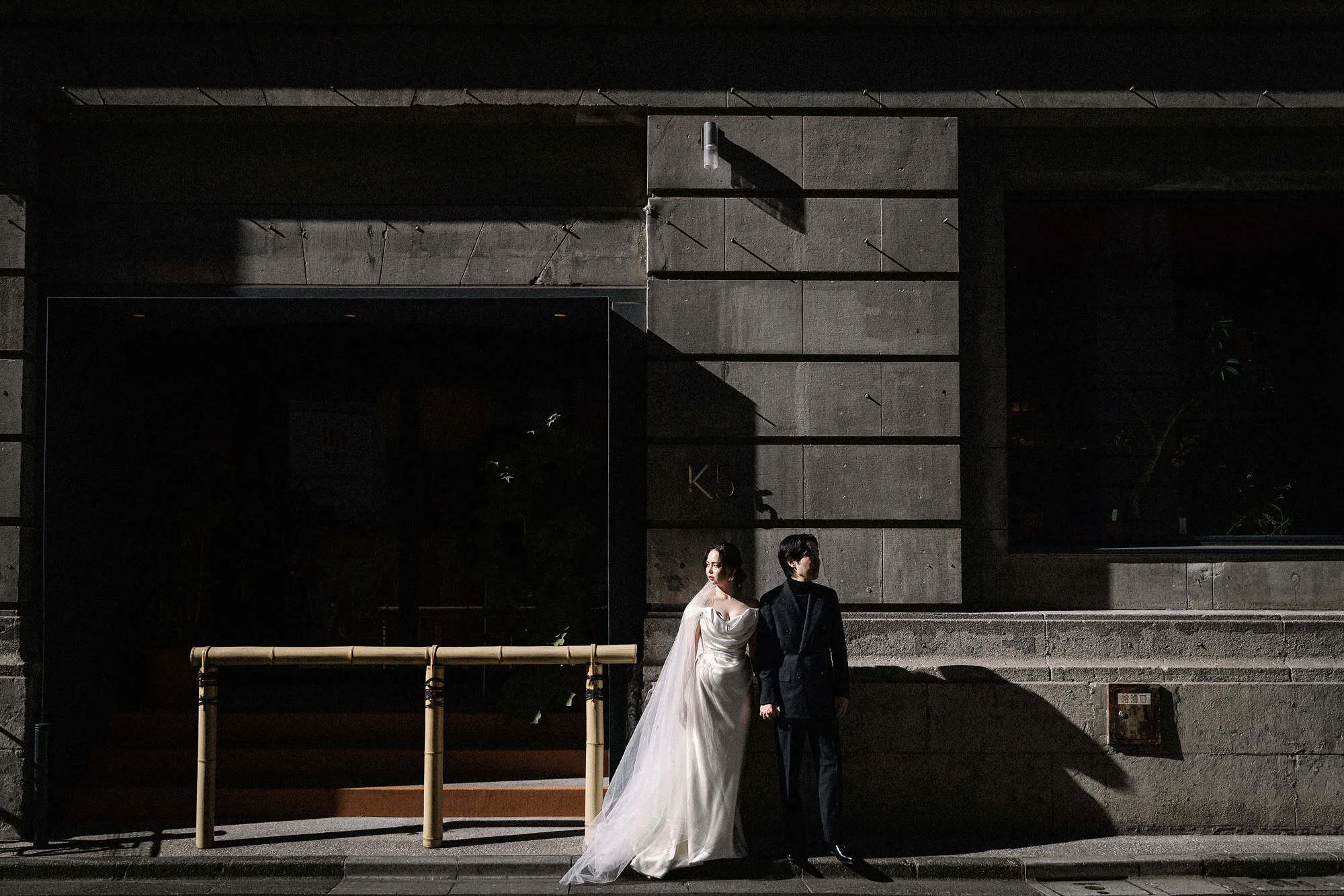 The bride and groom stand in a slice of hard sun against the dark stone of a 1923 bank building in Kabutocho on the day of their Tokyo Bay wedding.