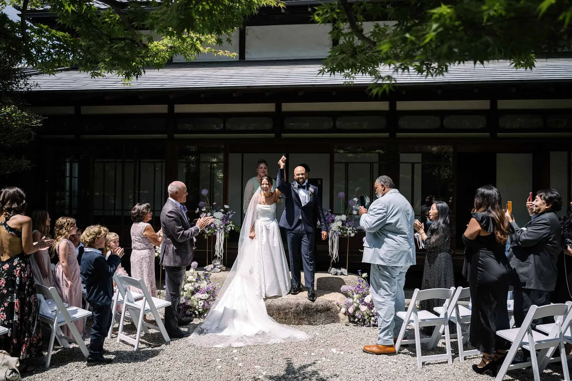 Family and friends cheer as the just married couple raise their joined hands at a Tokyo garden wedding, a traditional Japanese house behind them.
