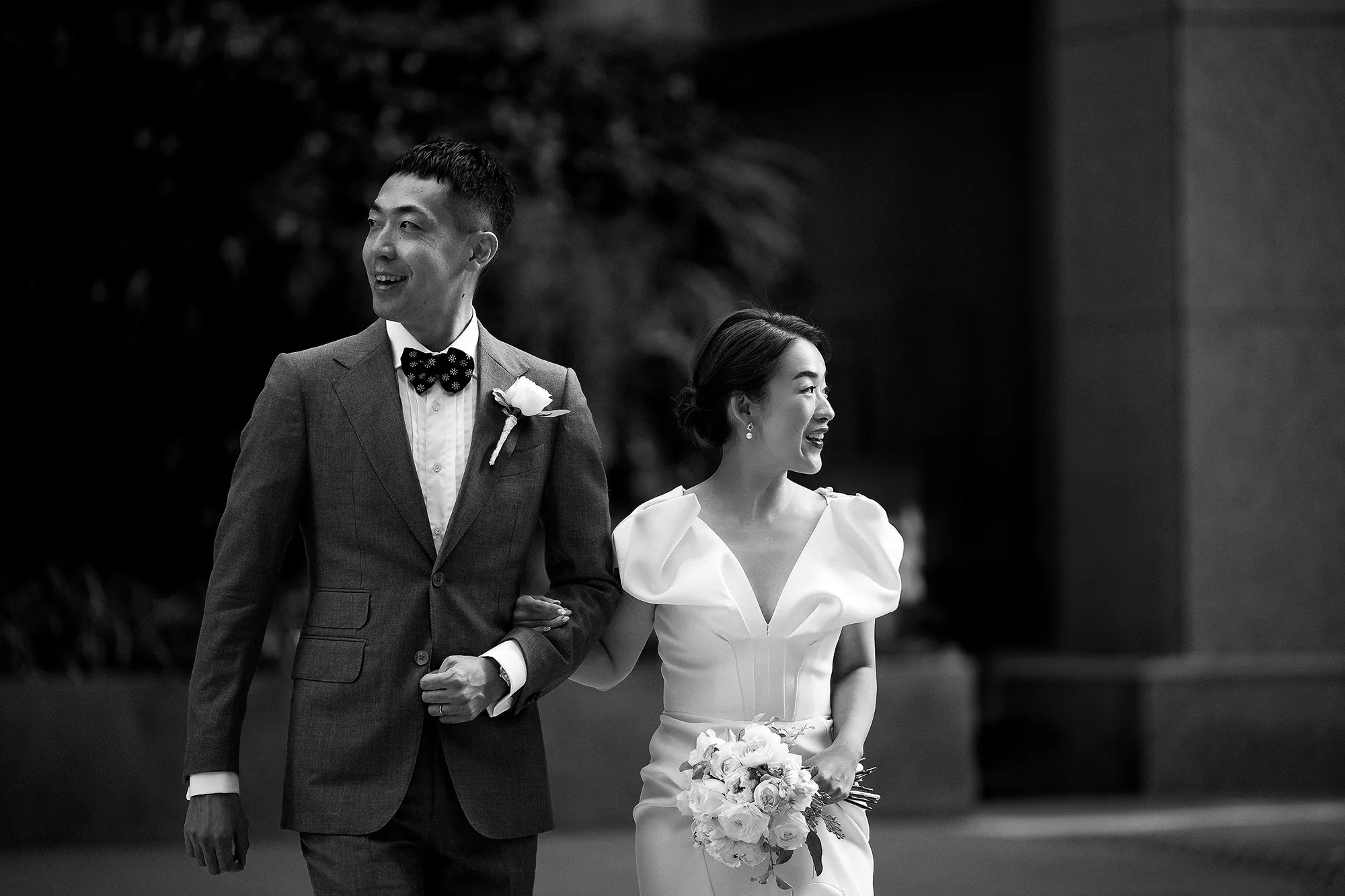 In black and white, the bride and groom walk arm in arm through the city, smiling, her bouquet of white roses in hand.
