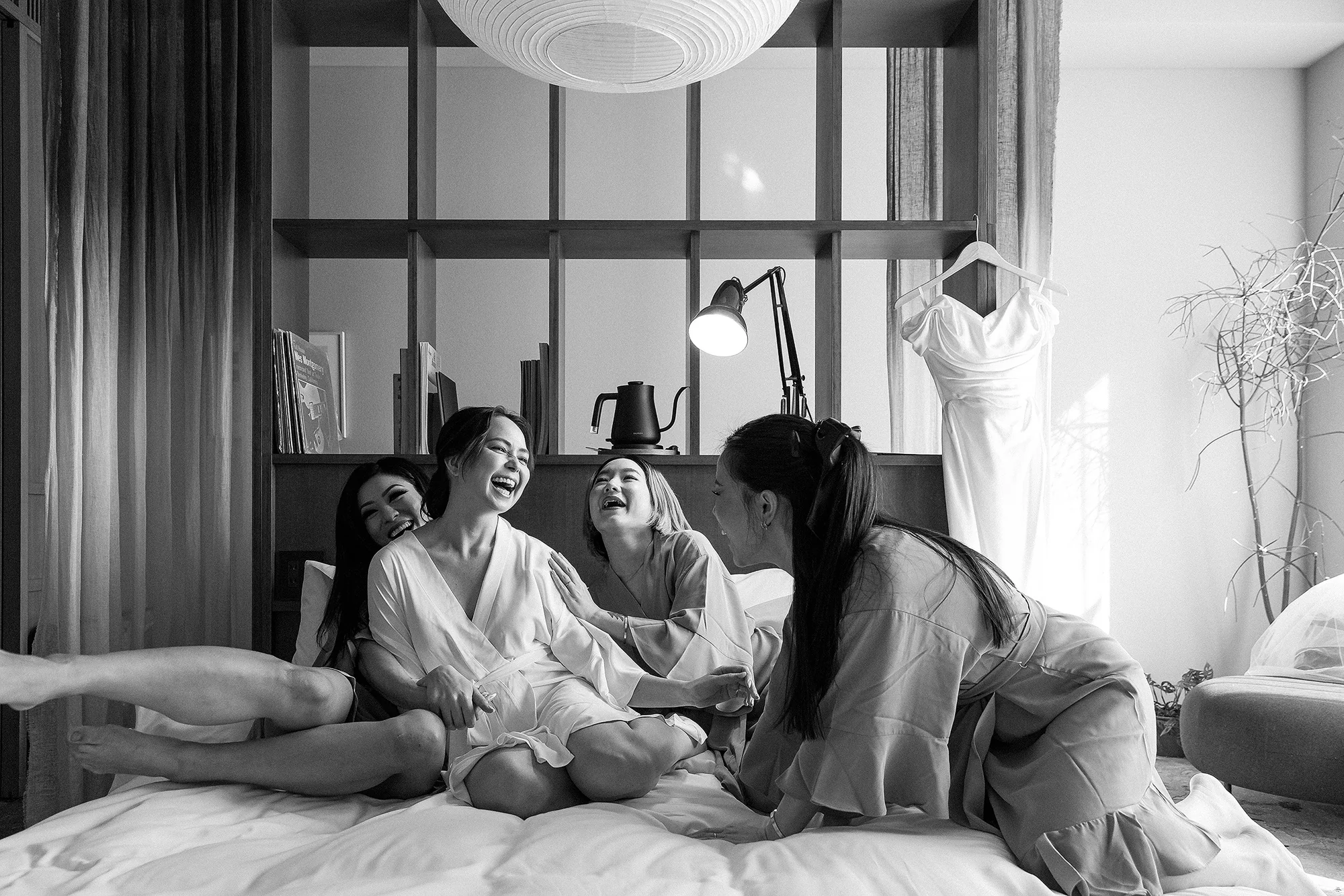 In black and white, the bride and her friends laugh together on the bed while getting ready in a Tokyo hotel room.