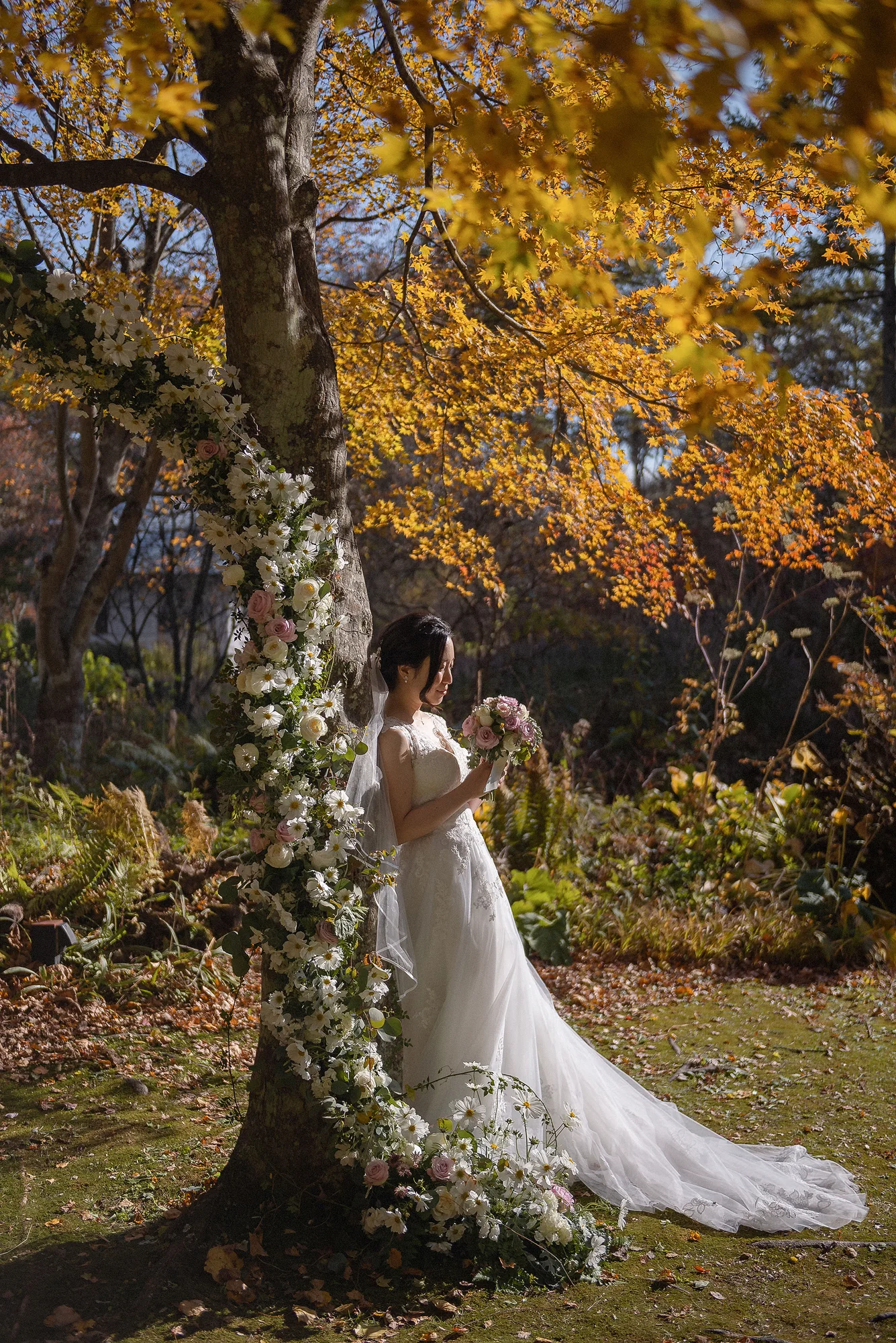 The bride stands at the foot of a golden maple, its trunk wound with white flowers, in an autumn garden in Karuizawa.
