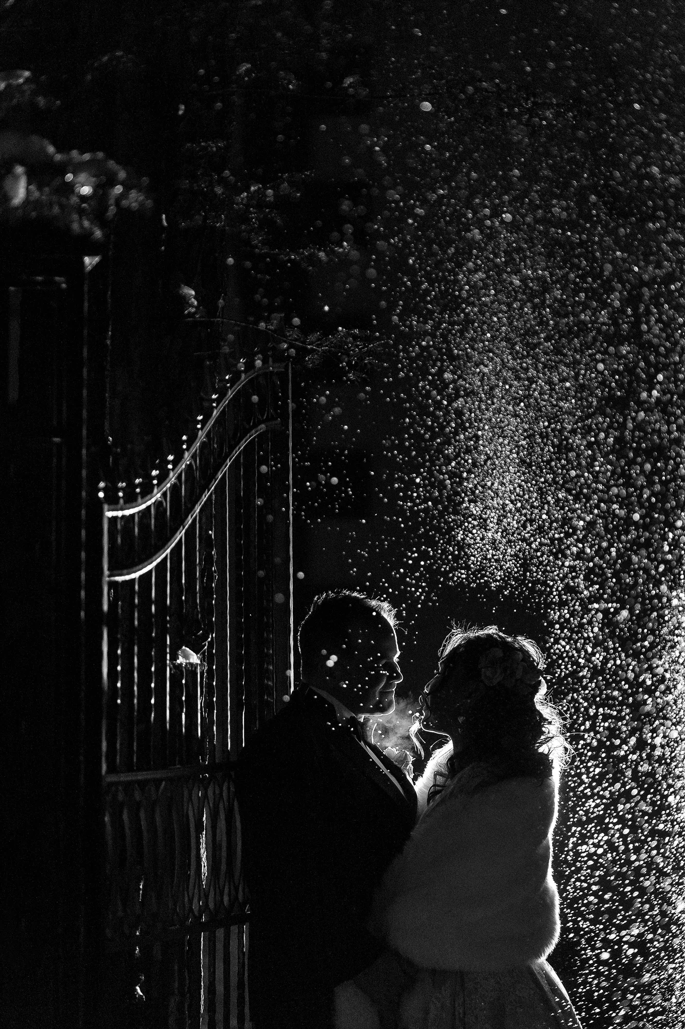 In black and white, the couple stand close by an iron gate at night as snow falls through the light, in Karuizawa.