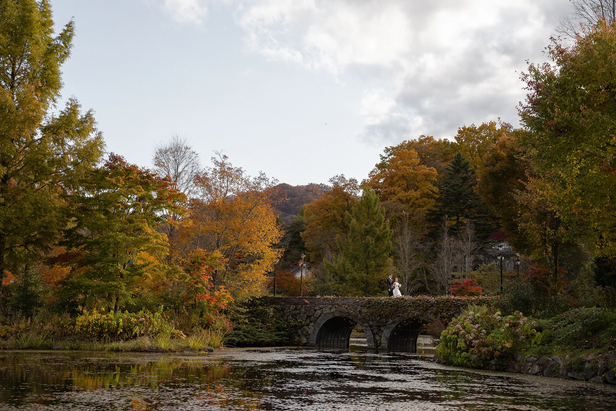 The couple stand small on an old stone bridge over a still pond, autumn trees all around, in Karuizawa.