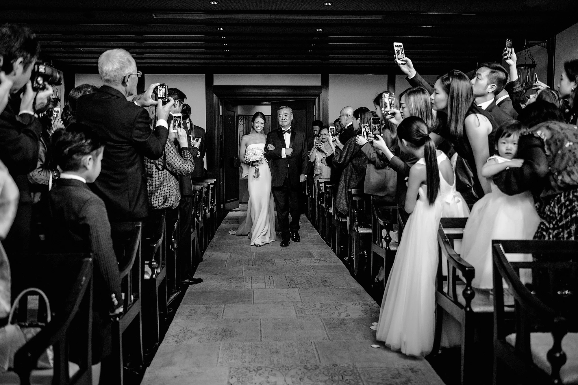 In black and white, the bride walks down the aisle with her father as guests hold up their phones.