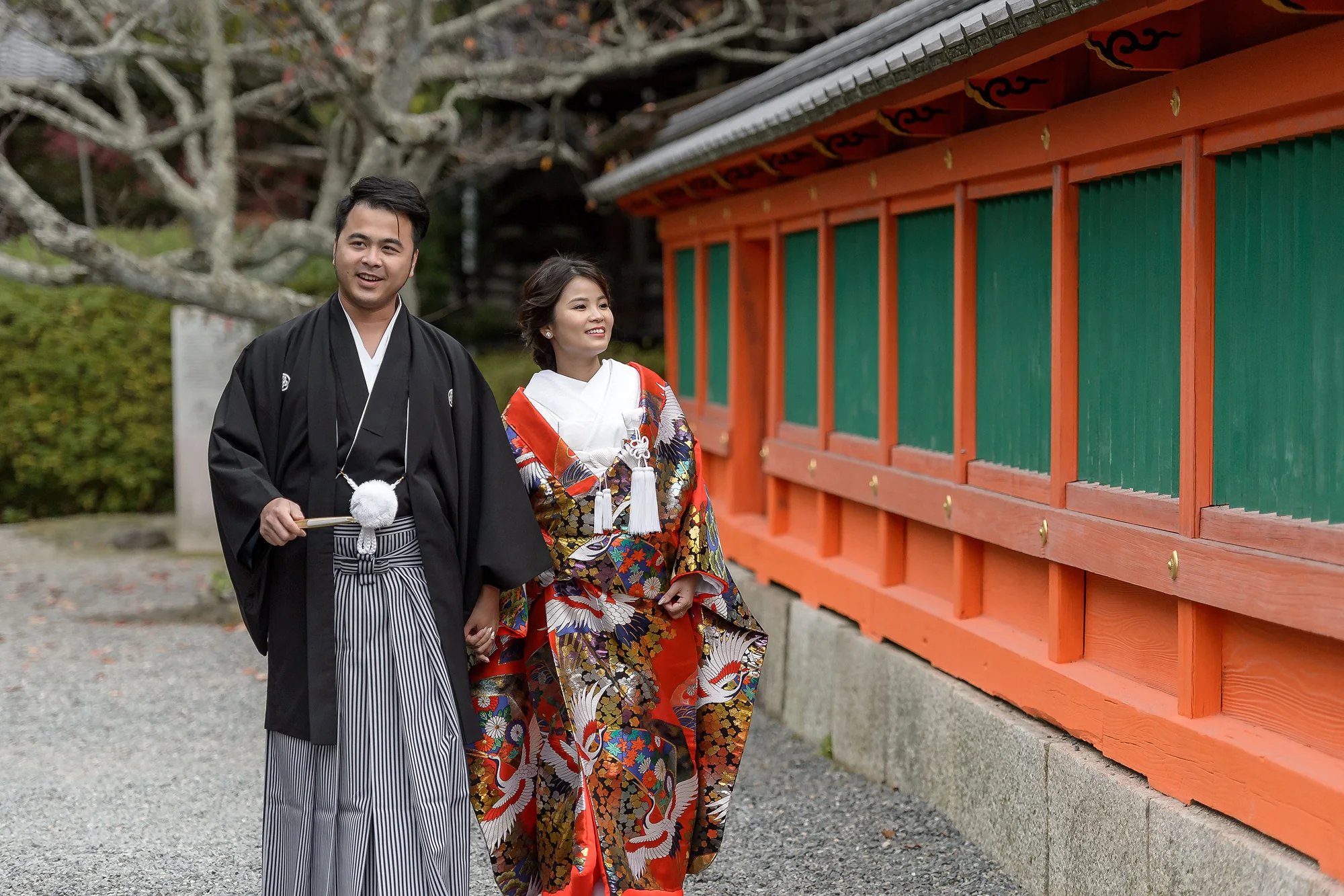 The couple in hakama and a red uchikake walk beside a vermilion shrine wall in Kyoto.
