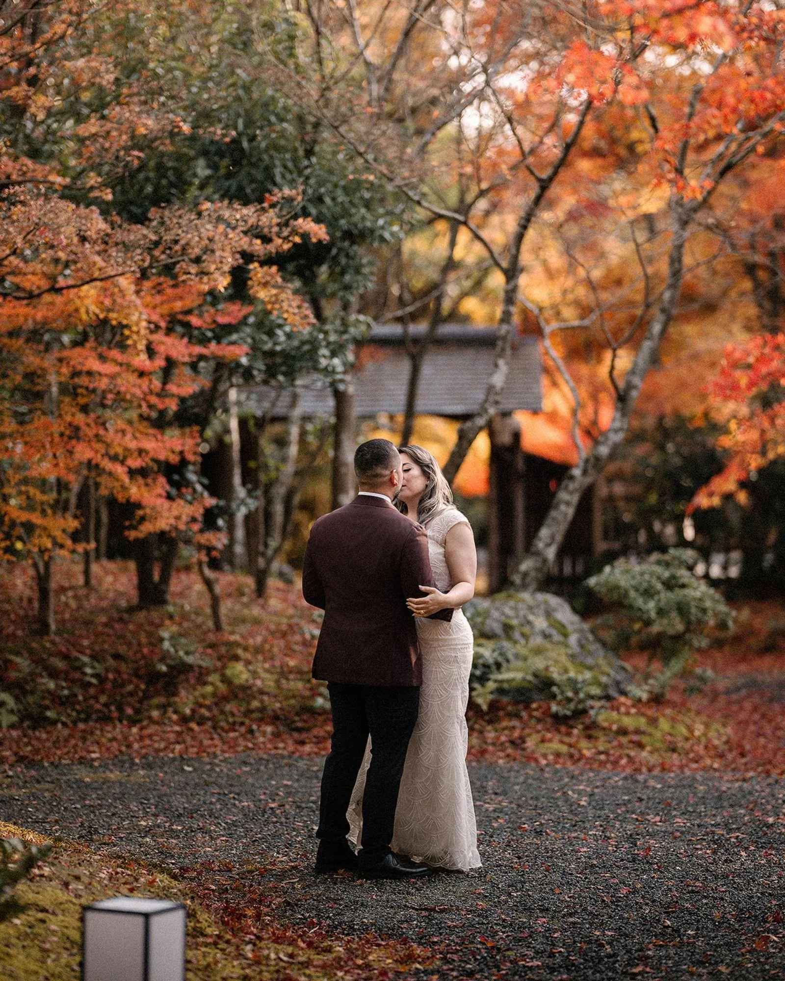The couple embrace beside a stone lantern in a Kyoto maple forest, leaves covering the ground.