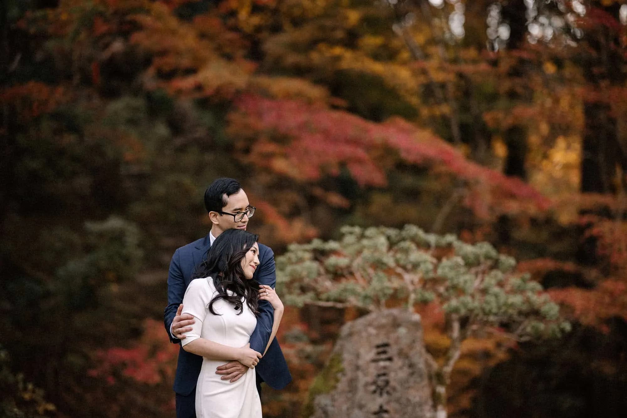 The groom holds the bride from behind in a Kyoto garden at peak autumn, red and orange maples and a carved stone marker around them.