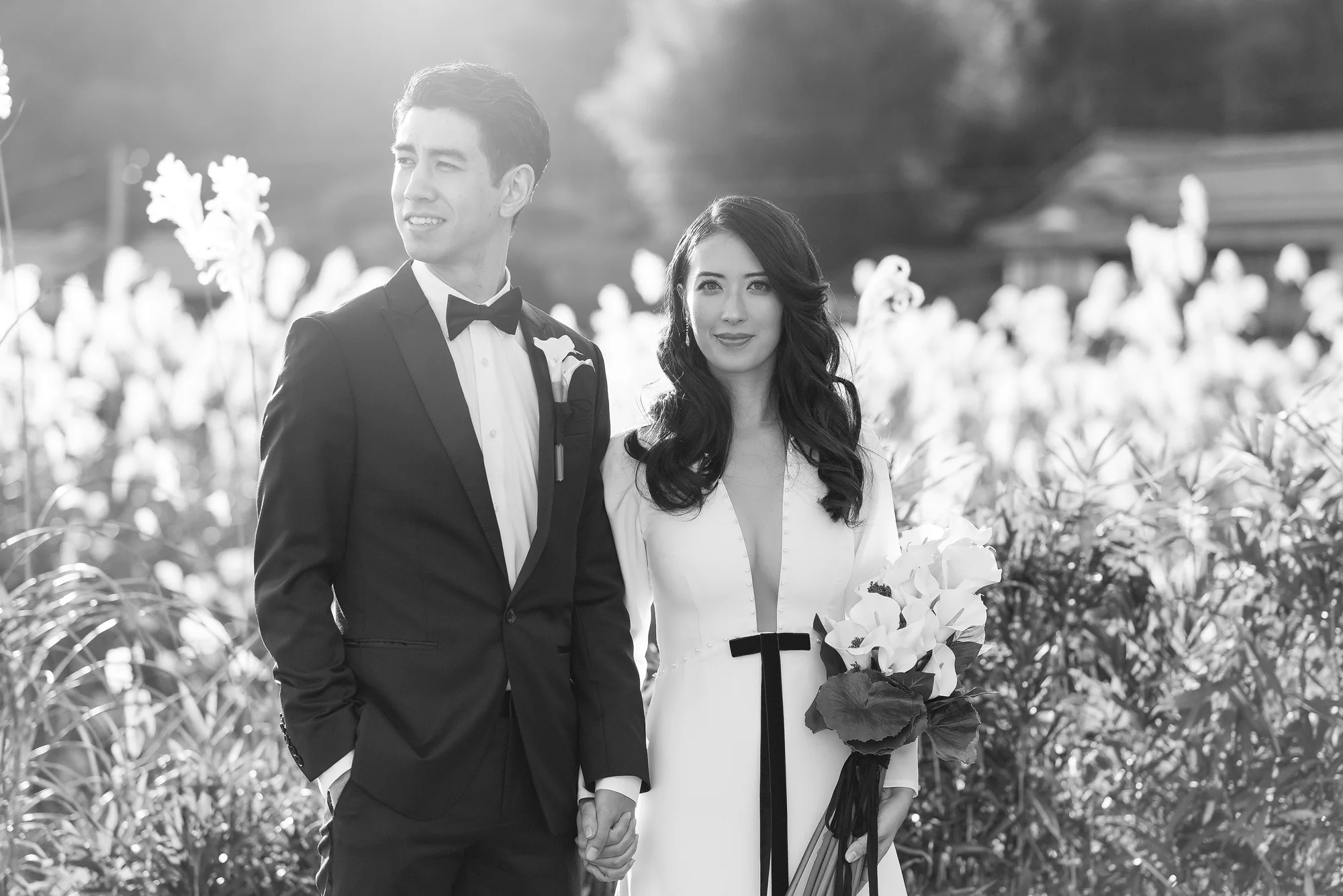 In black and white, the couple stand among pampas grass, the bride holding white lilies, in Kyoto.