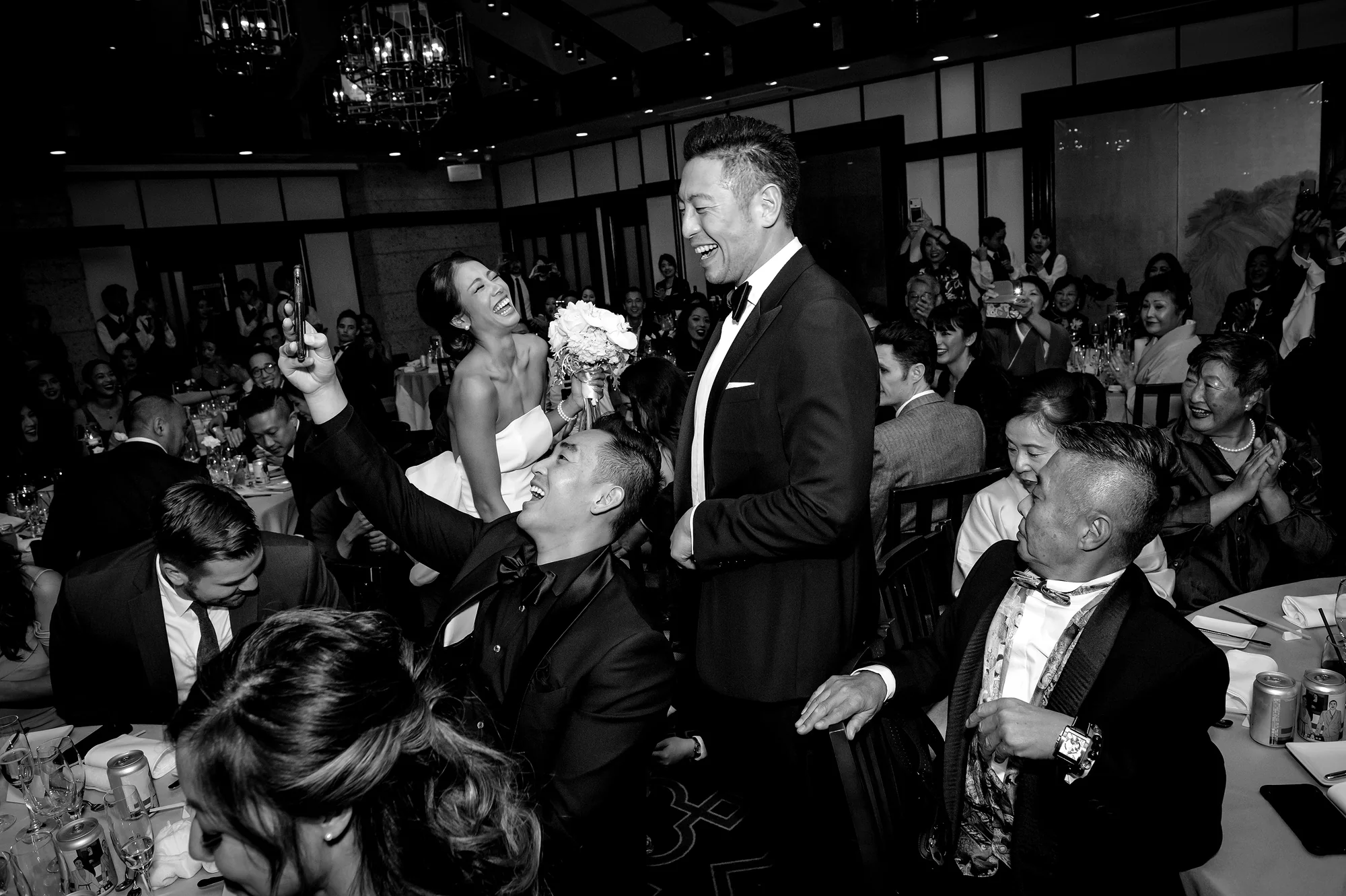In black and white, the couple walk into their reception laughing as guests cheer, film on their phones and reach out to them.
