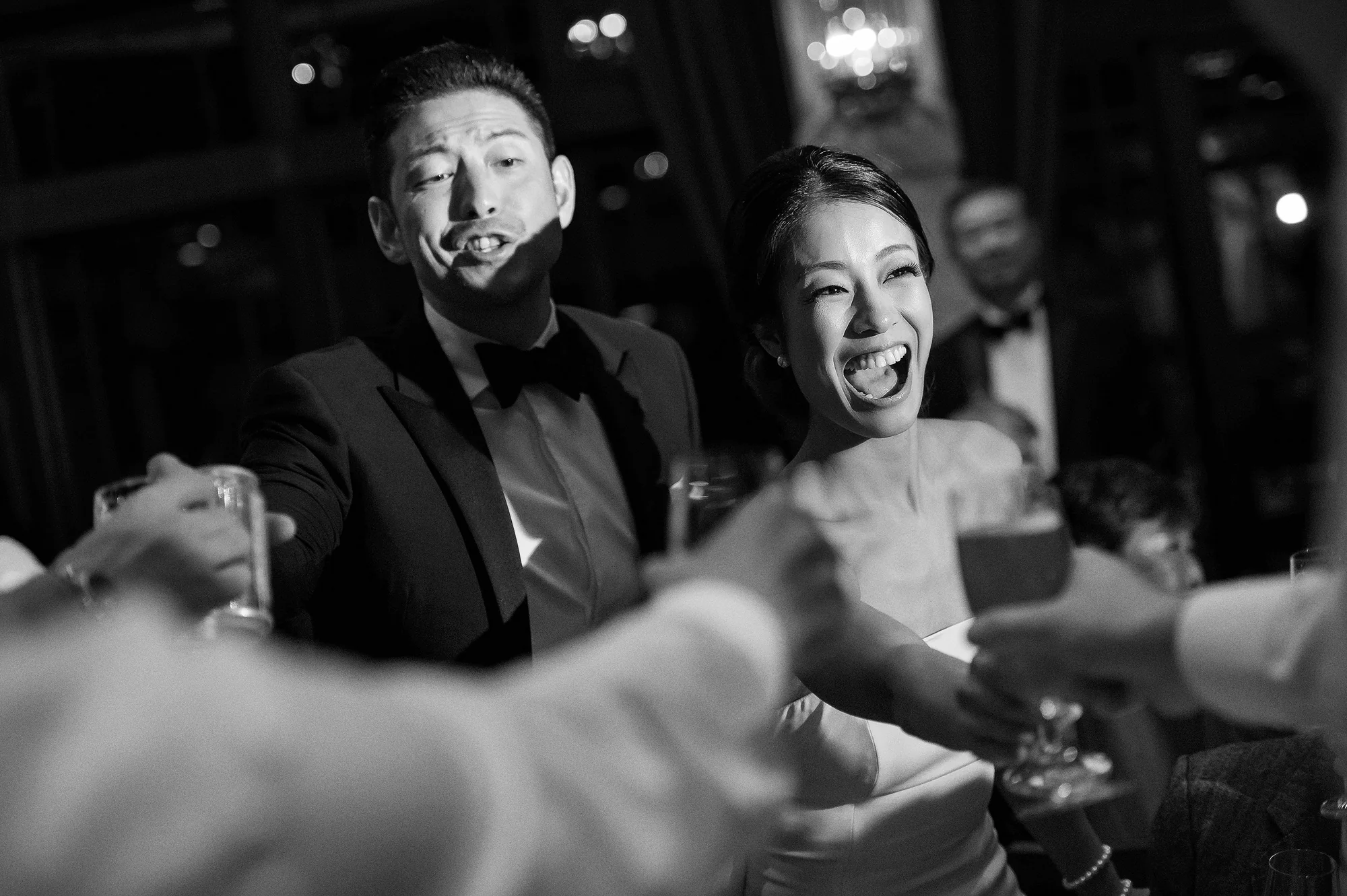 In black and white, the groom and bride laugh as guests raise their glasses around them in Kyoto.