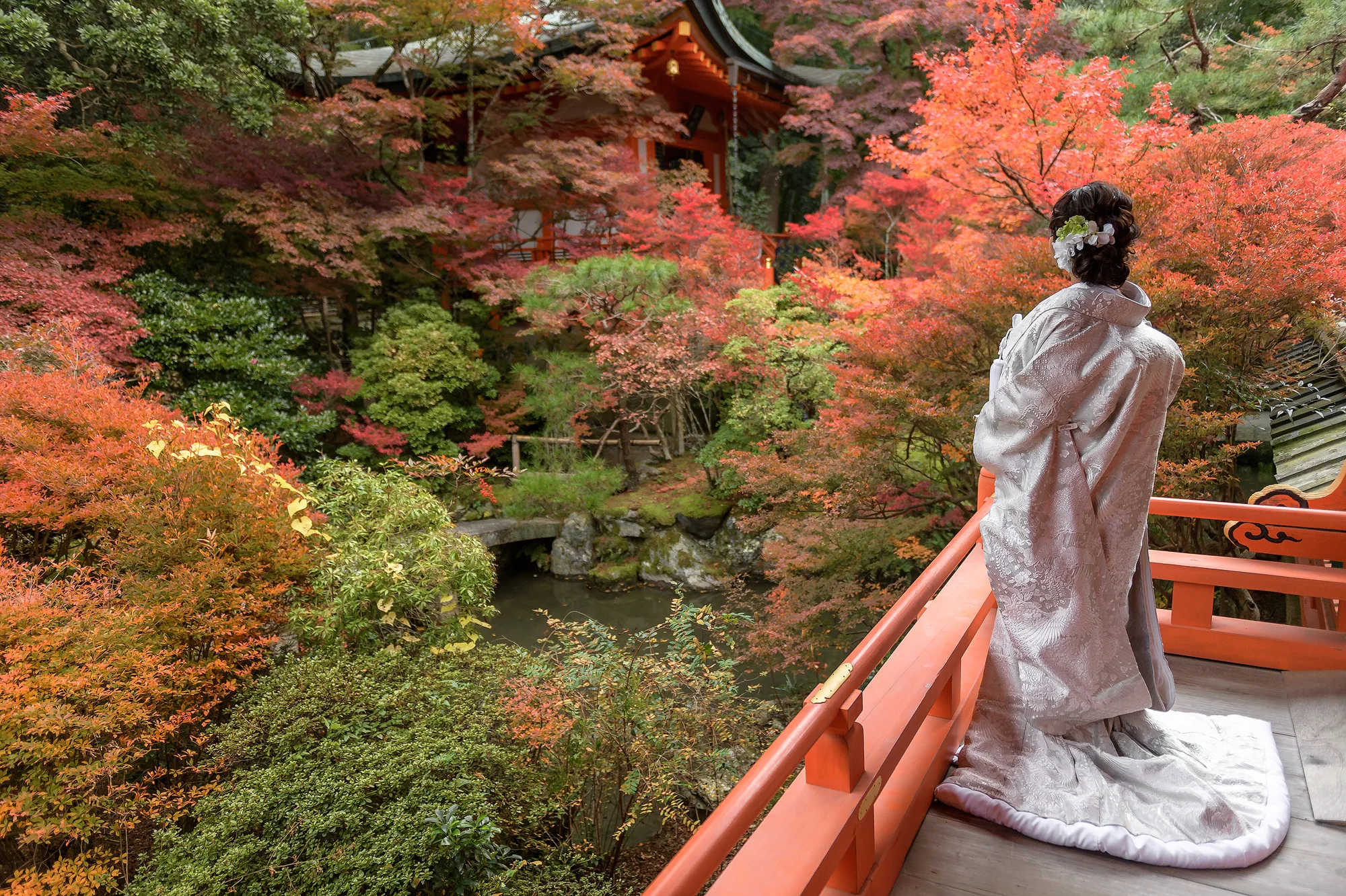 The bride in shiromuku stands on a vermilion balcony looking over an autumn garden in Kyoto.