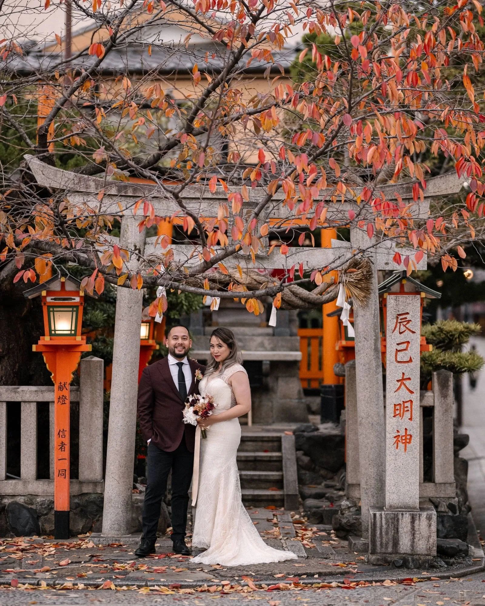 The couple stand beneath a stone torii hung with vermilion lanterns in Kyoto.