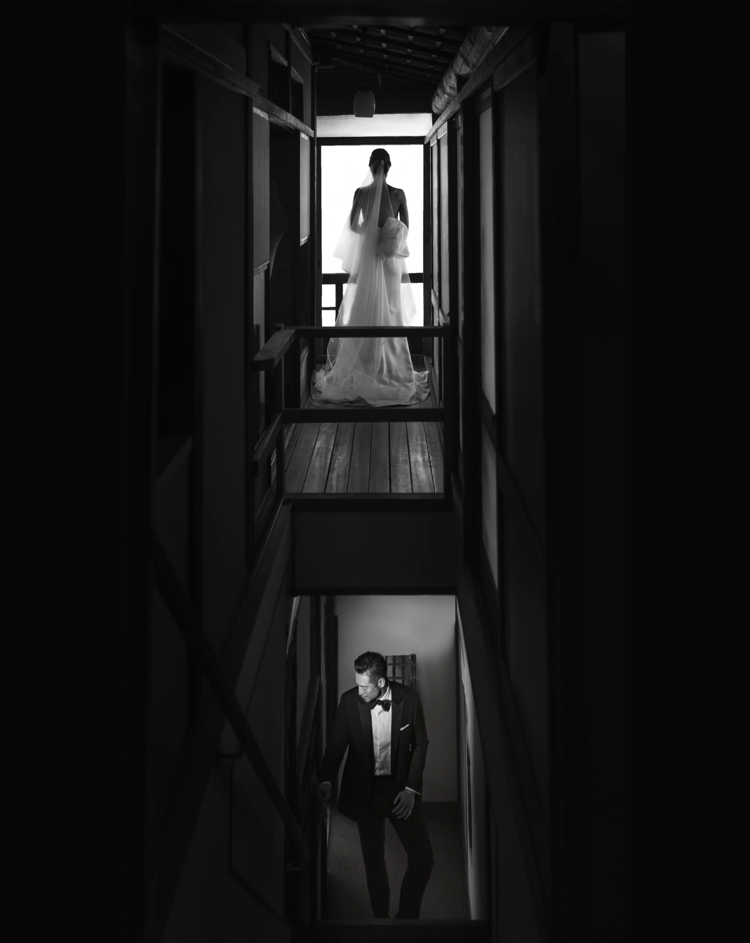 In a narrow dark stairwell, the bride waits at the top in the light as the groom stands below.