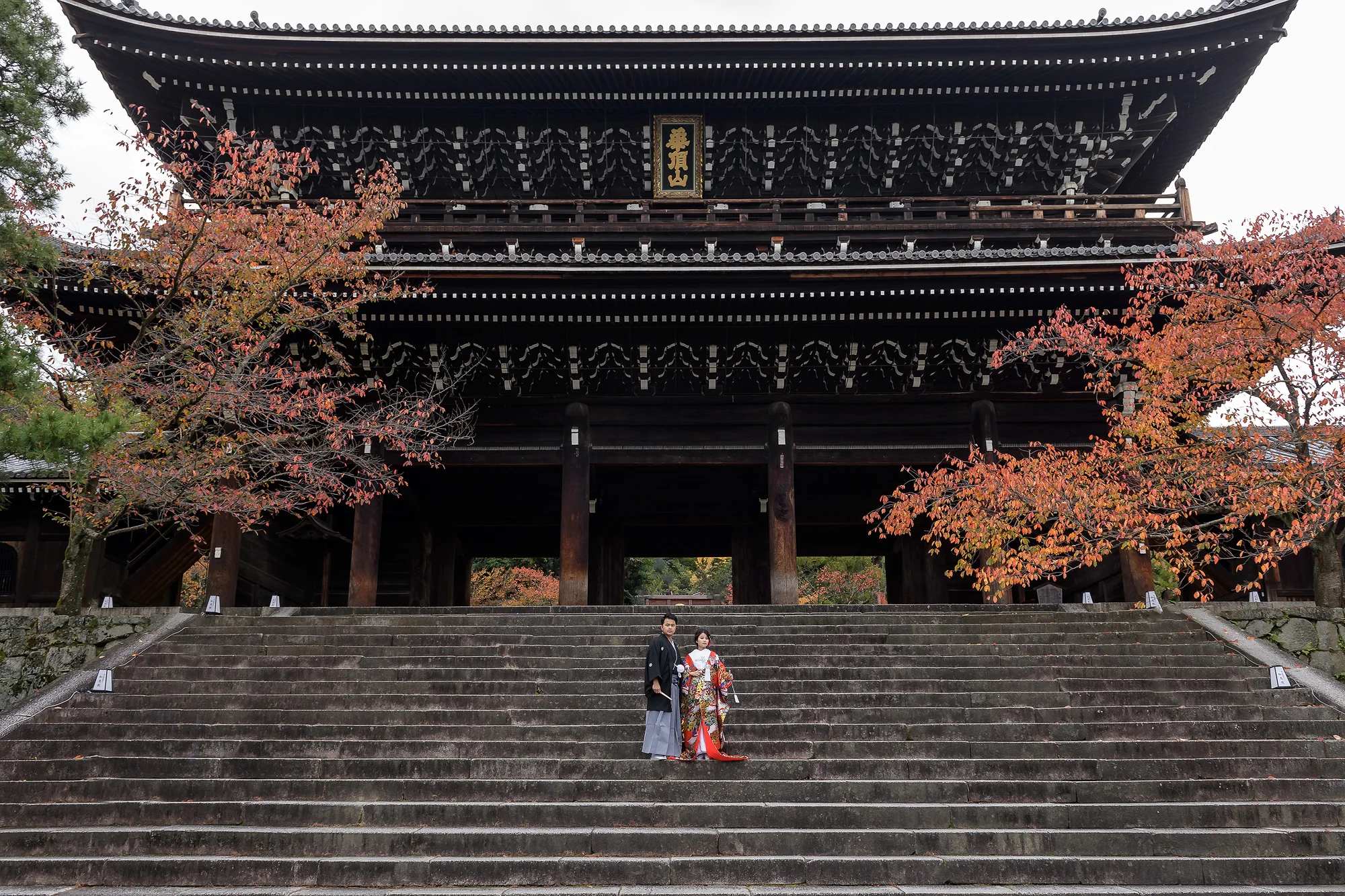 The couple in kimono stand on the wide stone steps below a great temple gate framed by autumn trees in Kyoto.