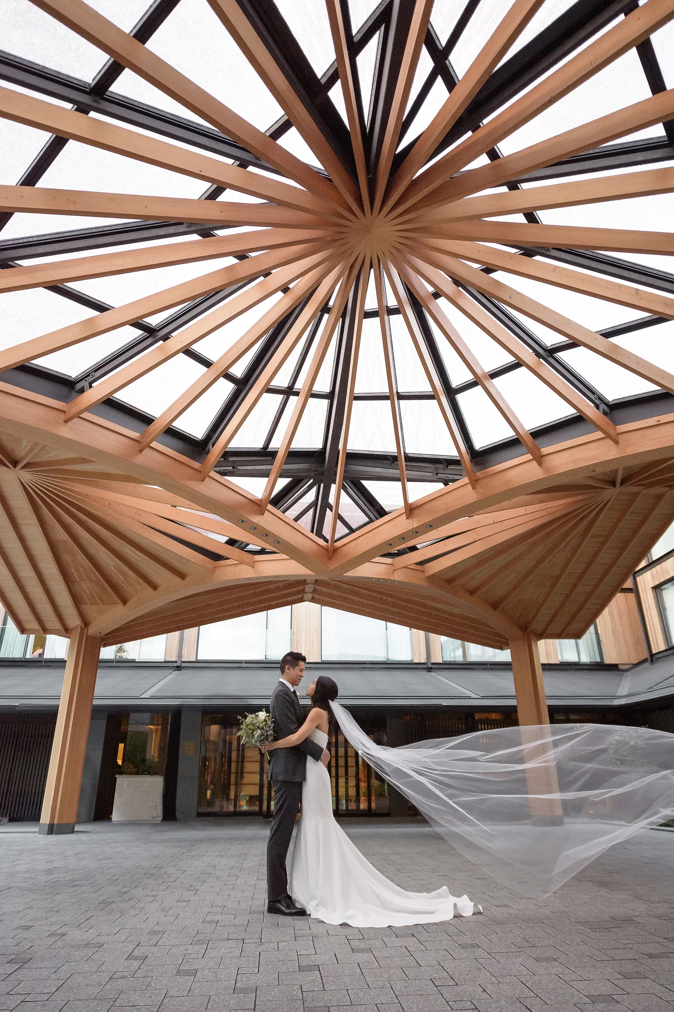 Beneath a radiating timber dome, the couple embrace as the bride's veil lifts across the courtyard in Kyoto.