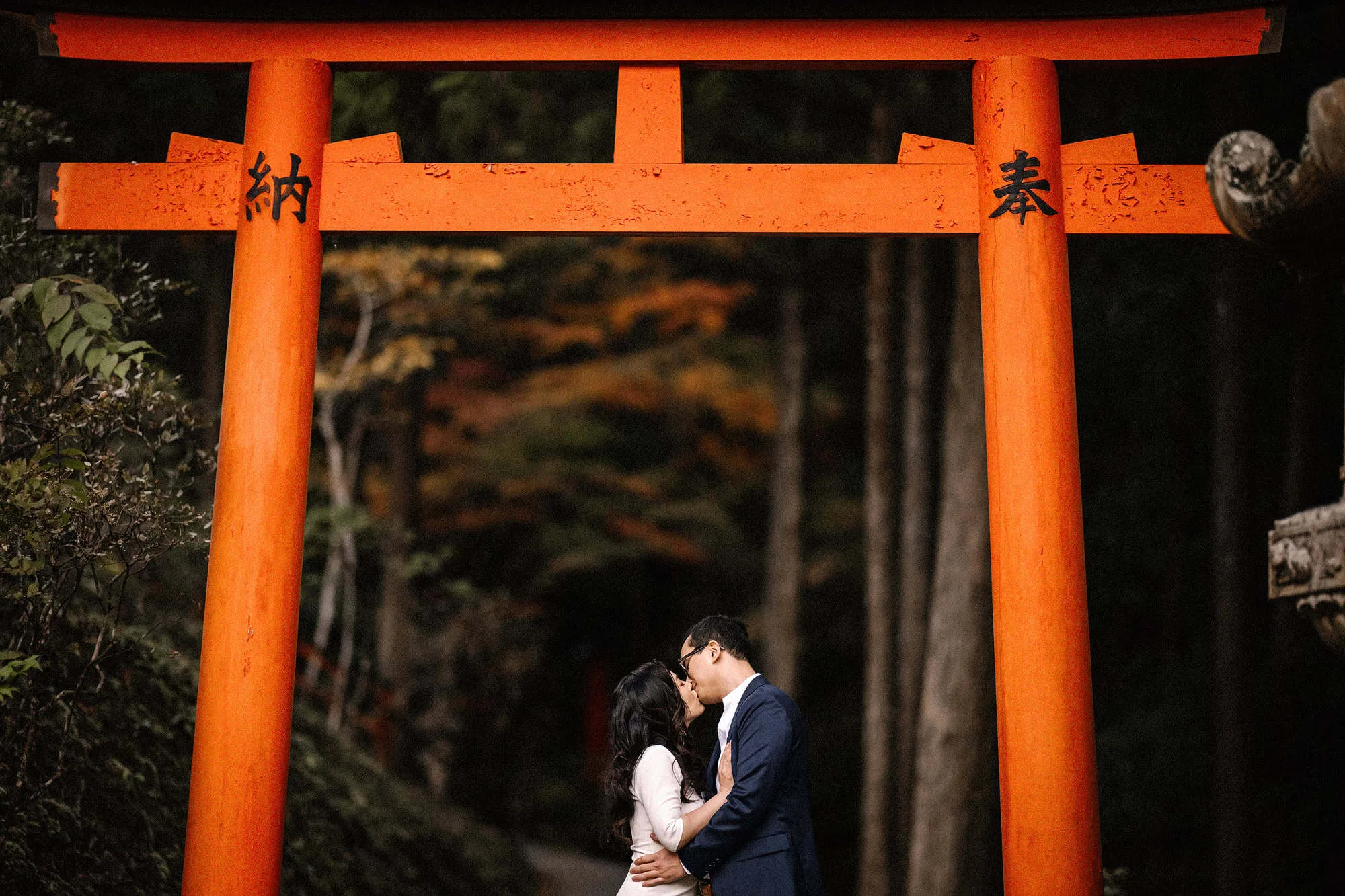 The couple kiss framed by the pillars of a vermilion torii, orange maples glowing in the forest behind, a Kyoto pre wedding shoot.